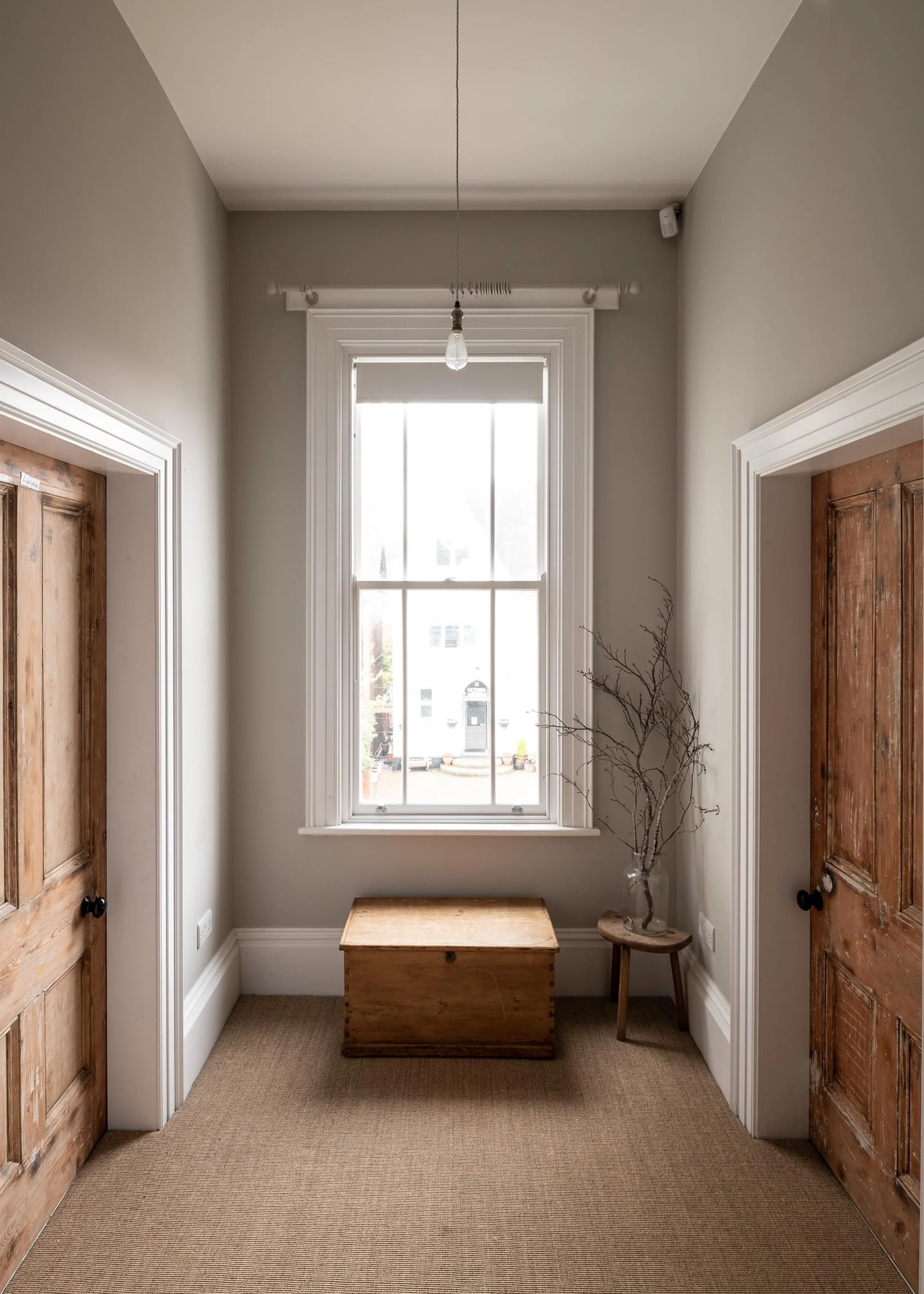 A small room with a window, a wooden trunk, a stool with a vase and some branches, and two wooden doors.