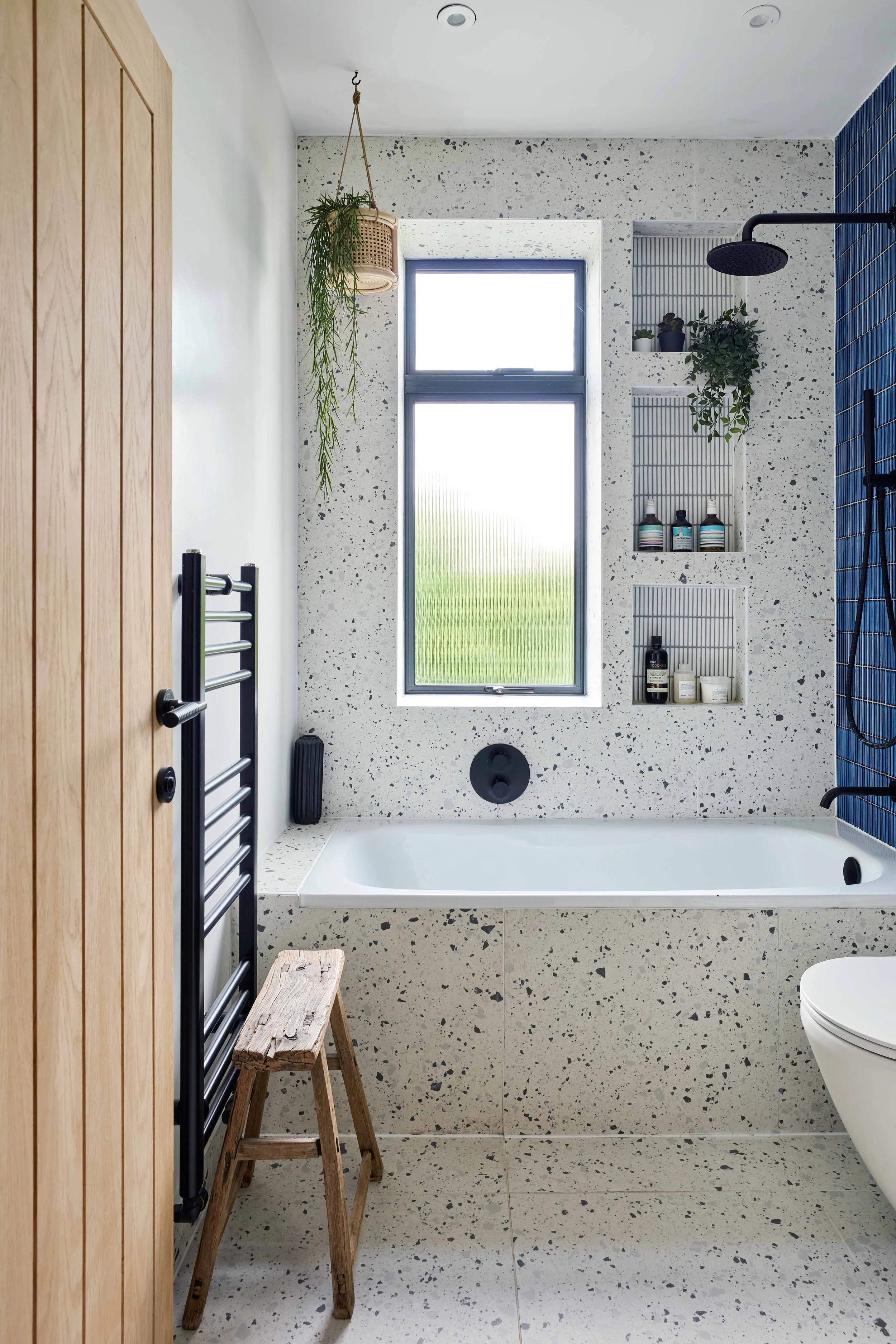 Modern bathroom with a terrazzo bathtub and wall, a window, built-in shelves with toiletries, a black showerhead, a towel radiator, a small wooden stool, and hanging plants.