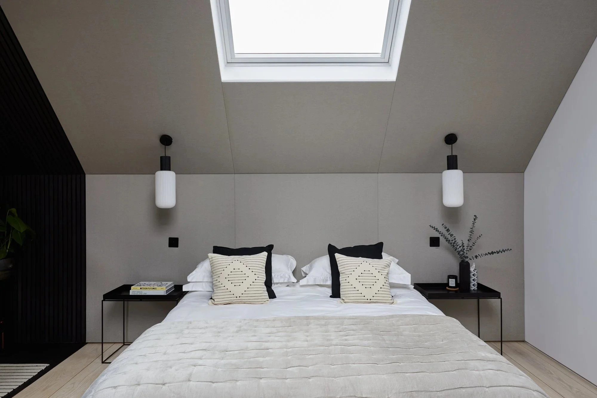 Modern bedroom with a skylight, two pendant lights, a bed with white and black pillows, black nightstands with books and decor, and a beige blanket.