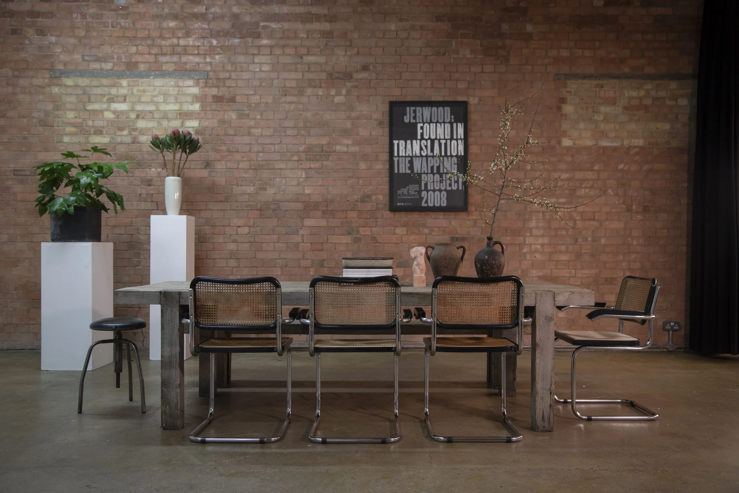 Interior of a room with a wooden table, six chairs, and decorative vases and plants against an exposed brick wall.