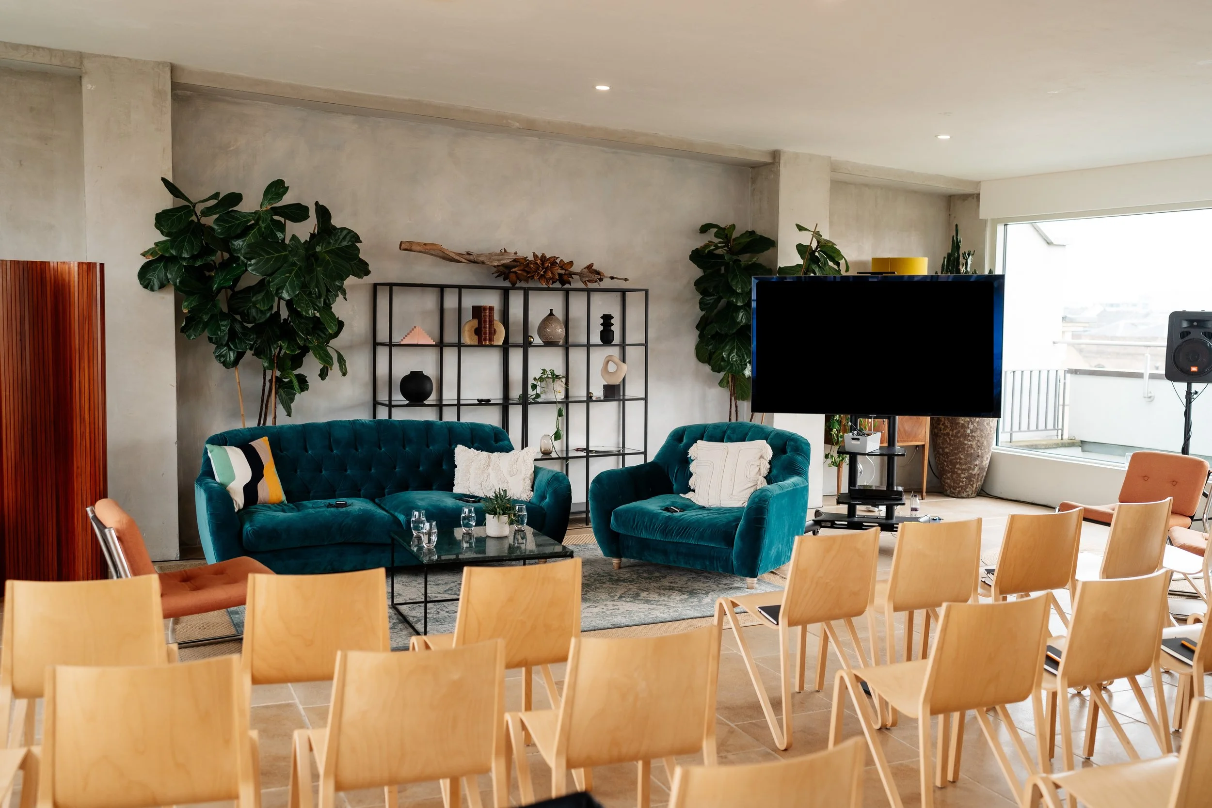 Living room with teal velvet sofas, a glass coffee table, wooden chairs, and large potted plants near the windows. There is a TV on a stand, a black metal bookshelf with decorative items, and a balcony in the background.