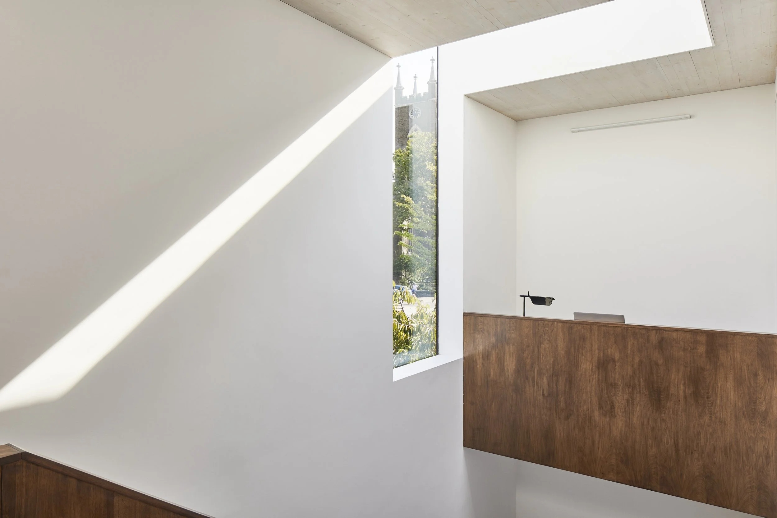 Modern interior space with white walls, angled ceiling, and a large vertical window showing a church tower outside, with sunlight casting a shadow inside.