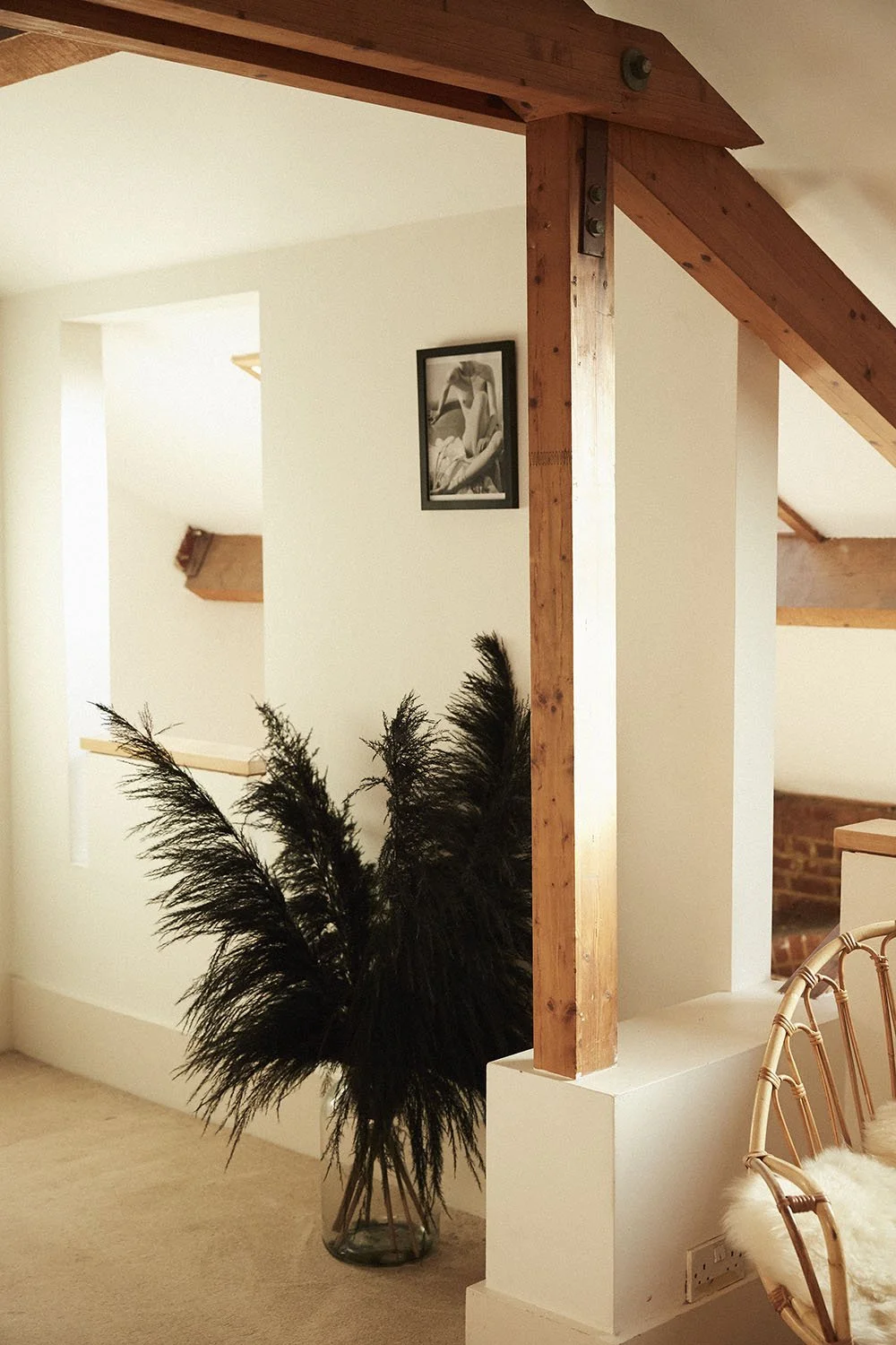 Interior of a house with a wooden beam, a potted plant with dark feathers, a black and white artwork on a white wall, a wicker chair with a white cushion, and exposed brick wall in the background.