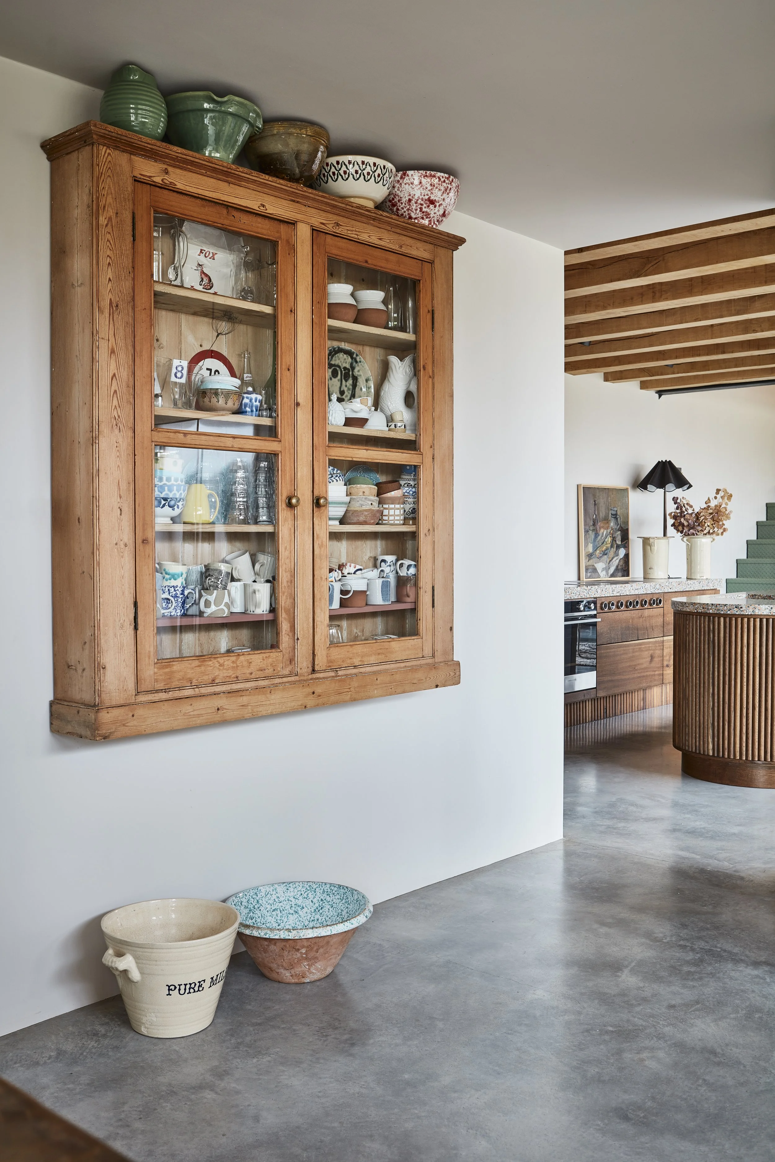 A wooden display cabinet filled with various dishes and glassware in a modern kitchen. Large decorative bowls are on top of the cabinet, and there's a countertop with a painting, vase, and dried flowers. The room features a wooden ceiling with expose