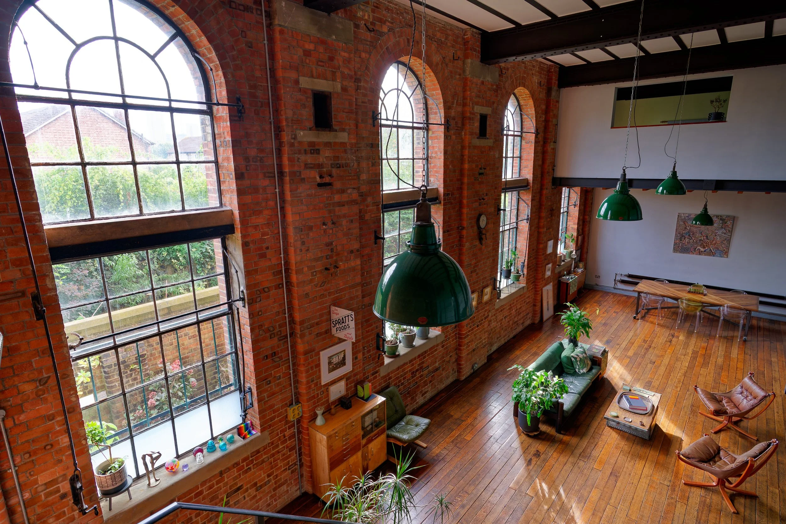 Interior of a spacious industrial-style loft with large arched windows, brick walls, wooden floors, green pendant lights, a green sofa, two brown chairs, and plants.