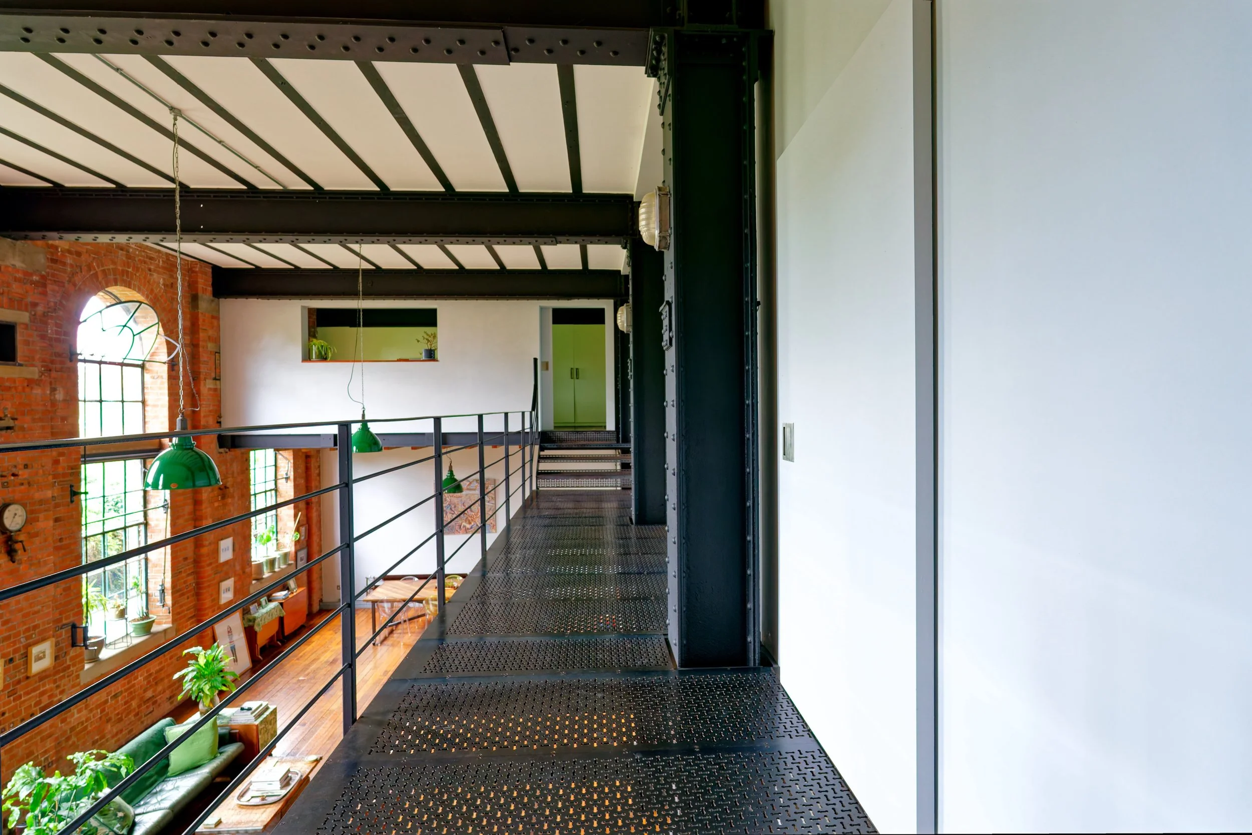 Interior view of a loft with black metal walkways, exposed brick walls, large windows, green hanging lamps, and indoor plants.