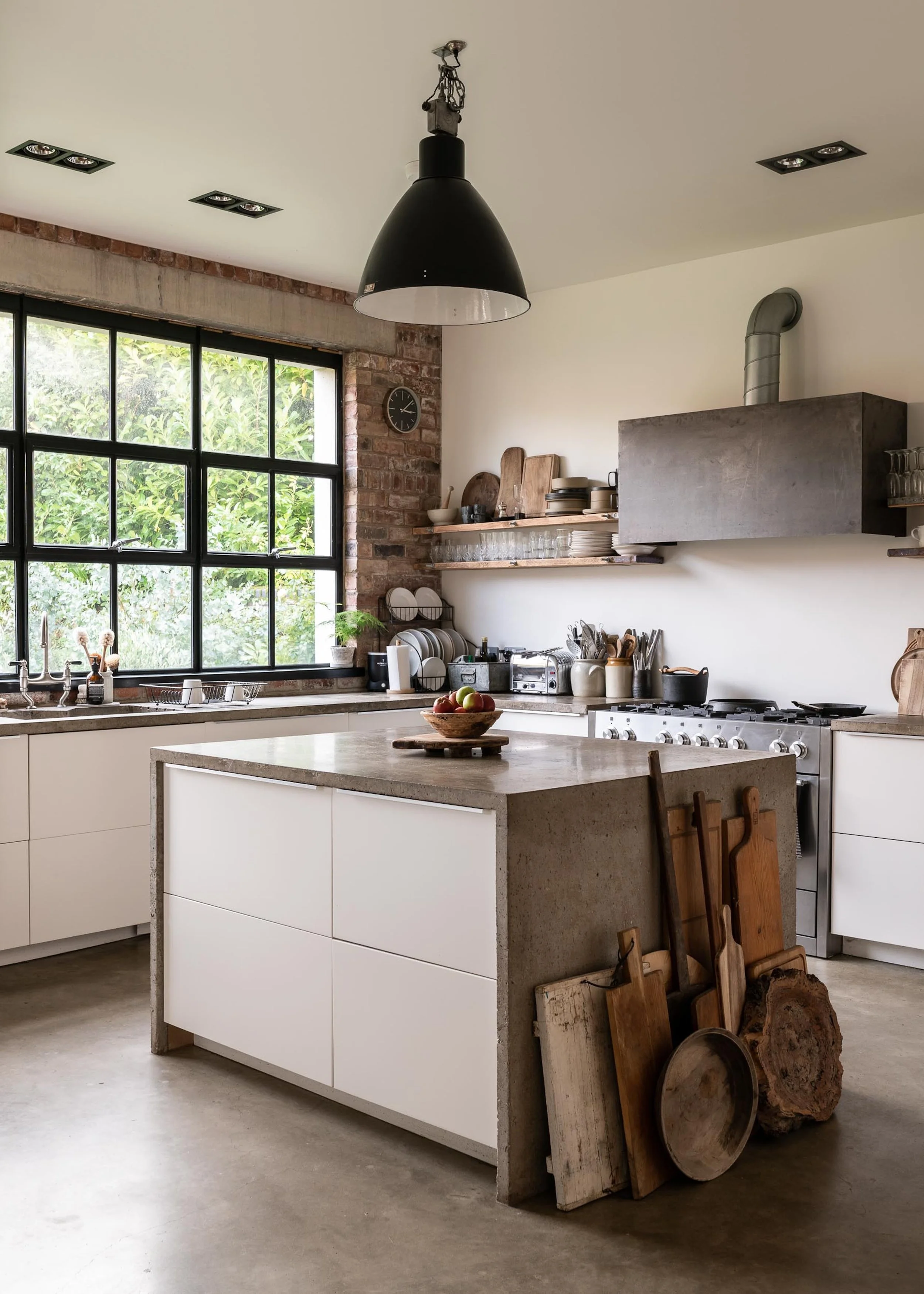 Modern kitchen with white cabinets, concrete countertop island, large window, exposed brick wall, and wooden cutting boards leaning against the island. Ceiling light fixture and various kitchen appliances and dishes are visible.