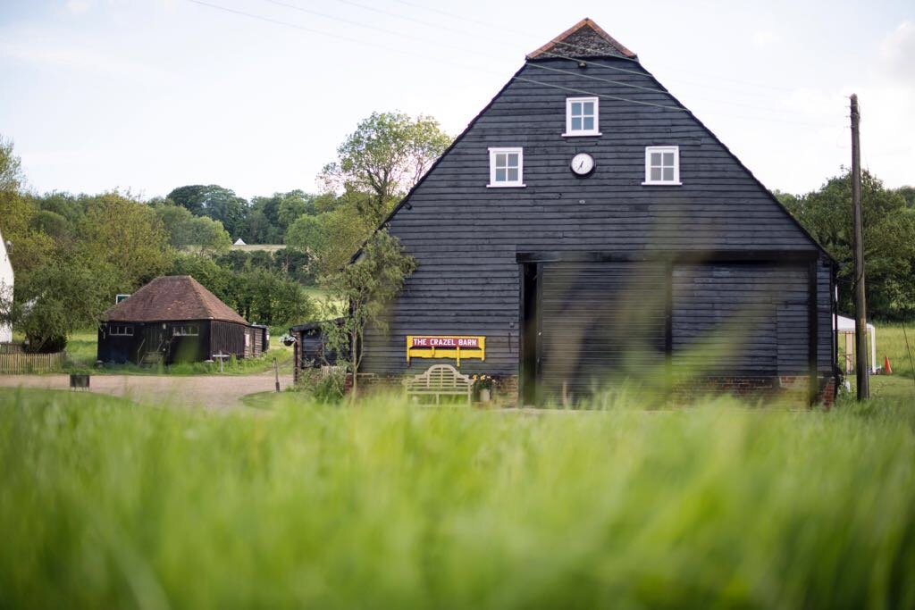 Black wooden barn with a clock and four windows on the upper level, set in a green rural landscape, with smaller black building in the background, and lush grass in the foreground.