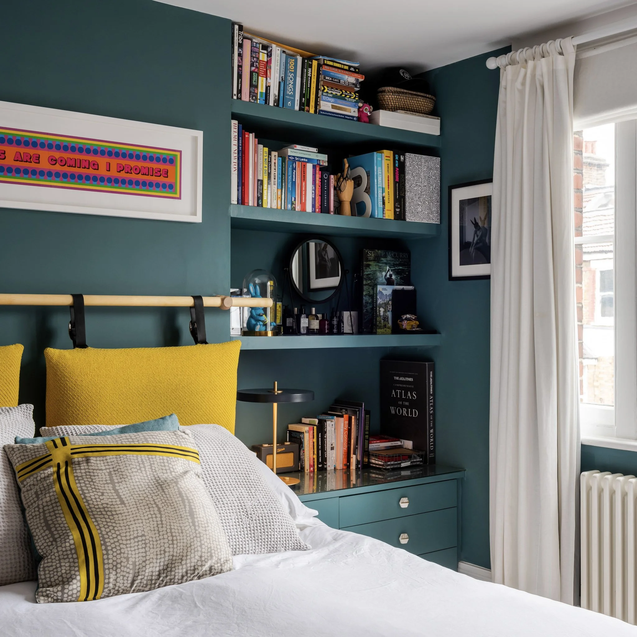 A bedroom with a teal wall, a bed with yellow and patterned pillows, a built-in shelf with books, a small round mirror, and framed artwork next to a window with white curtains.