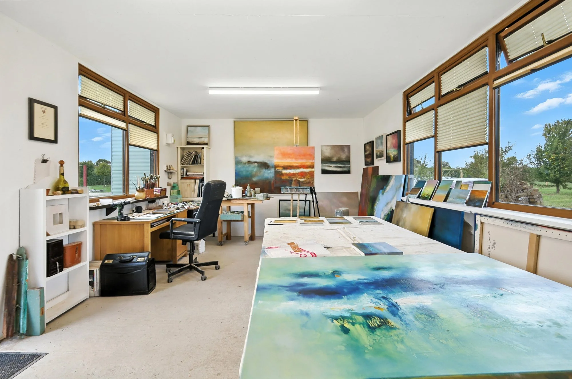 Artist's studio with large windows, paintings, art supplies, and a work table with painted canvases.