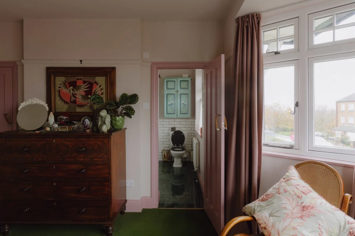 A cozy living room with a large window and pink curtains, a wicker chair with floral pillow, a wooden dresser with decorative items, and a view of a neighborhood through the window. In the background, a small toilet area is visible through an open do