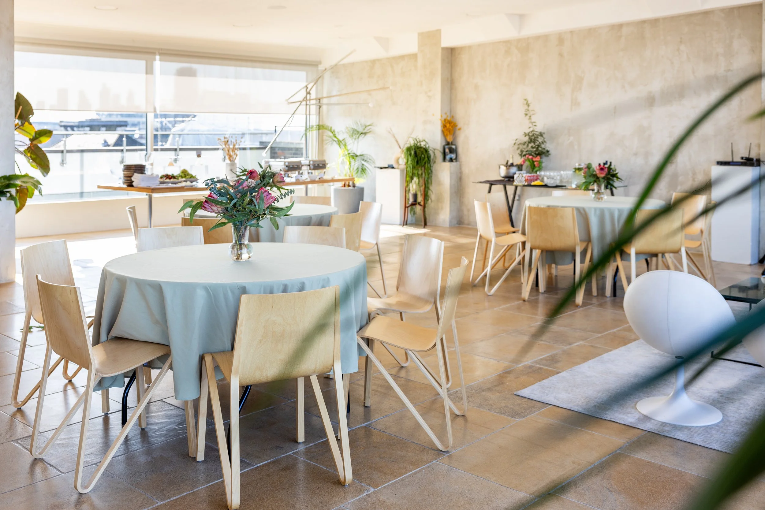 A bright, modern dining room with round tables draped in white tablecloths, decorated with floral centerpieces. Wooden chairs surround the tables, and large windows let in natural light. There are additional tables with snacks and flowers, and potted