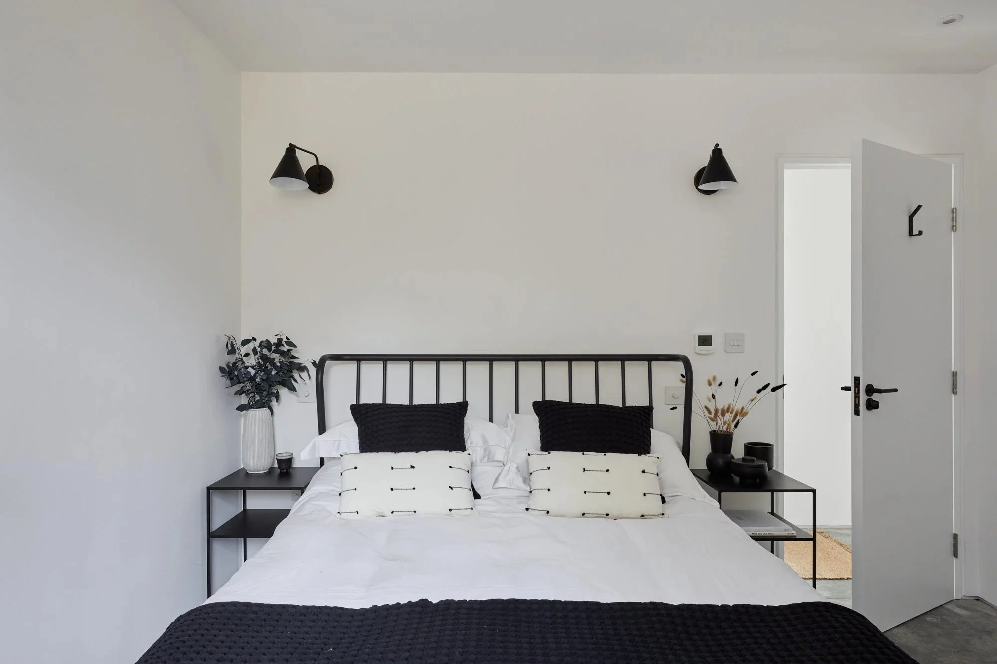 Minimalist bedroom with white walls, black bed frame, white and black bedding, black pillows, two black nightstands with decorative vases and plants, wall-mounted black lamps, slightly open white door.