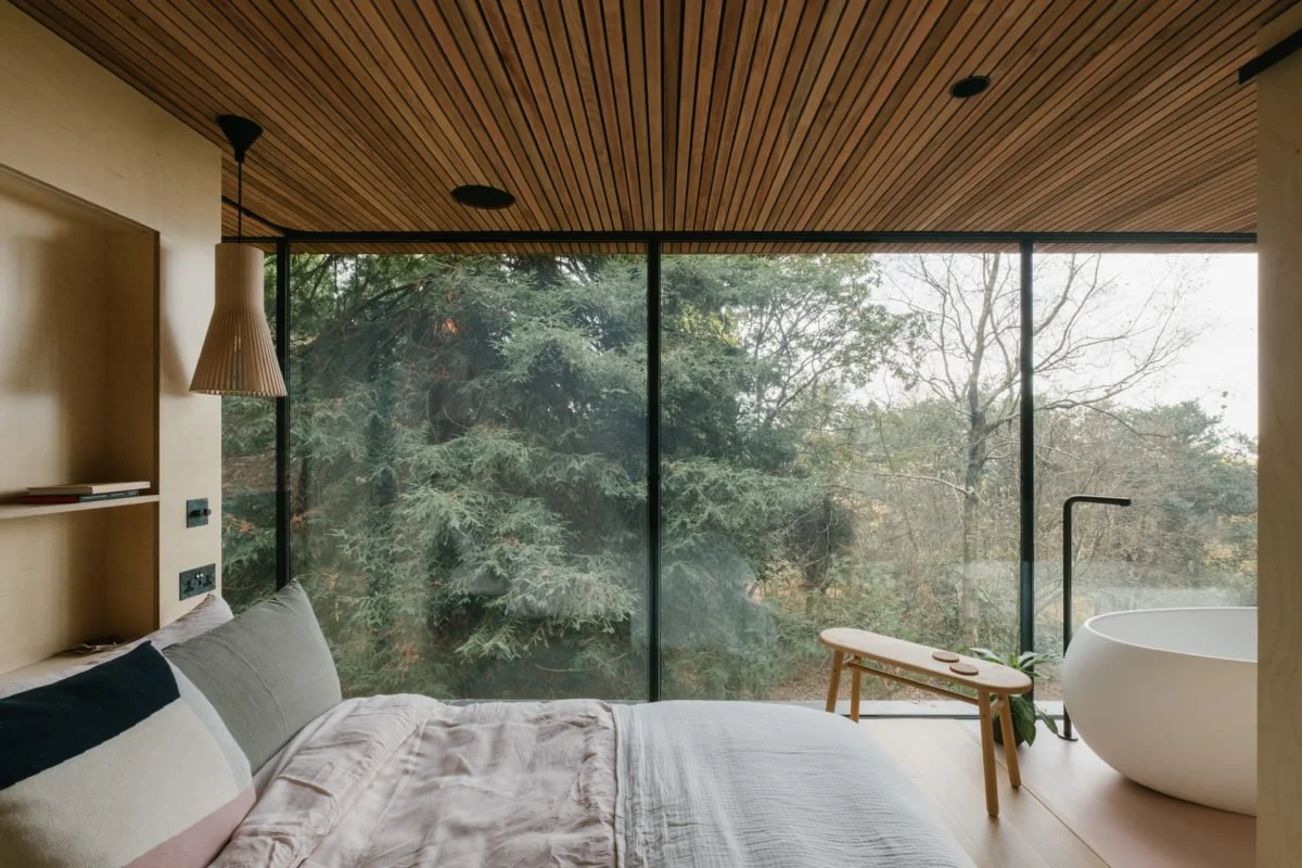 Modern bedroom with large glass window overlooking trees, with a bed, a wooden bench, and a standalone bathtub in the room.