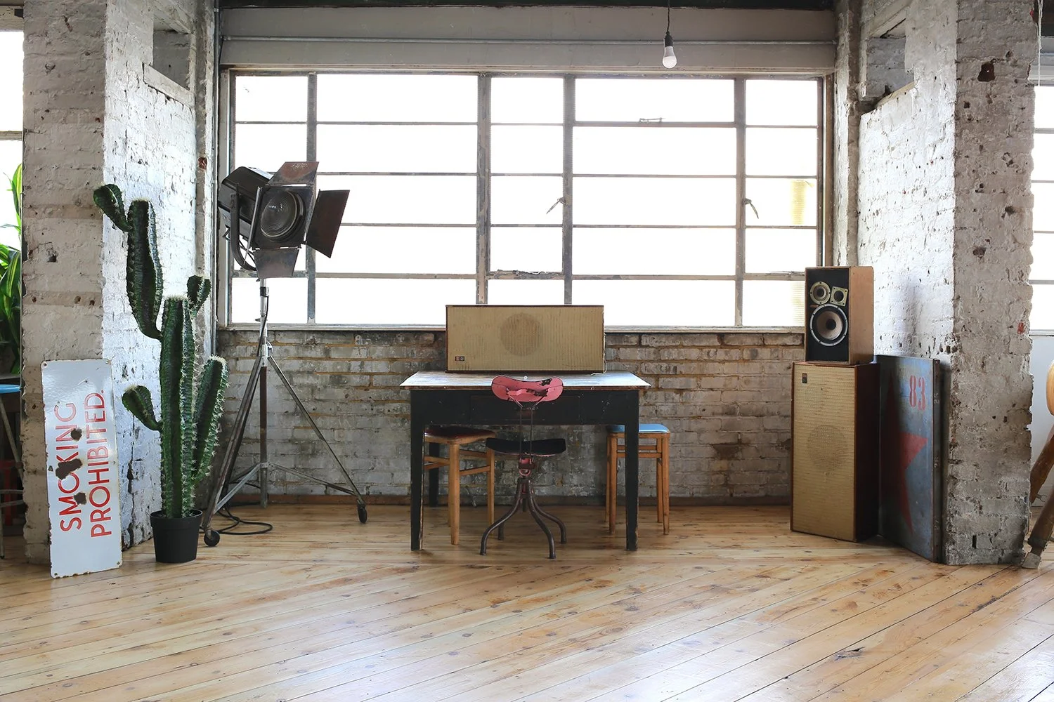 Interior of a room with brick walls, wooden floor, large window, vintage speakers on stands and a table, a cactus plant, a sign that reads 'Smoking Prohibited', and a pink bicycle seat attached to a stool.