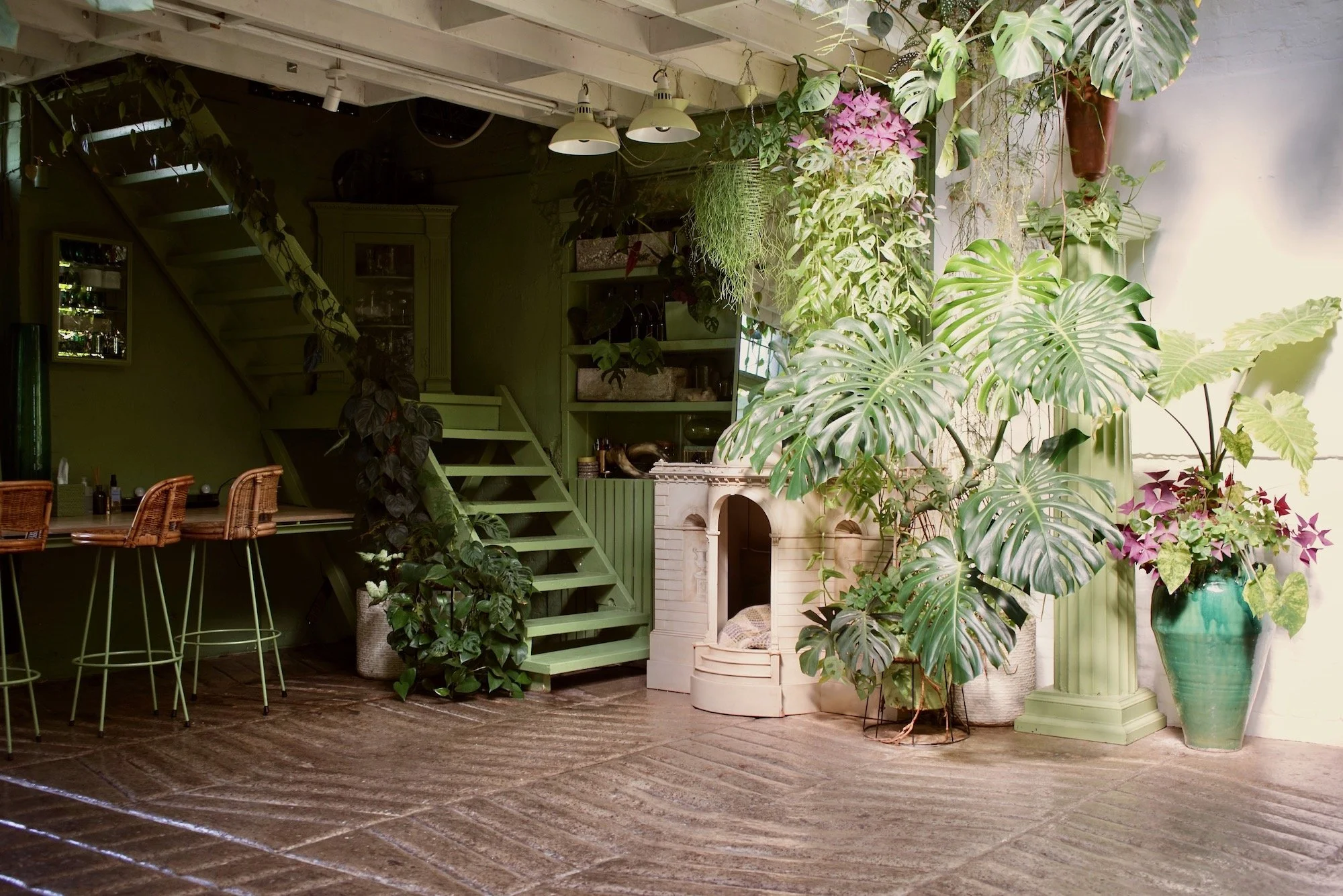 Interior of a room with green walls, wooden staircase, and a bar area with high chairs. The space is decorated with various green plants and a white fireplace. Bright natural light illuminates the space.