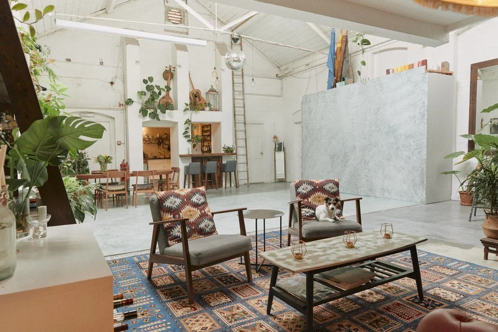 A spacious, well-lit indoor room with a bohemian decor style. It features comfortable armchairs with patterned cushions, a wooden coffee table with small candles, and a colorful rug. There are several potted plants, a white wall with wall-mounted gui