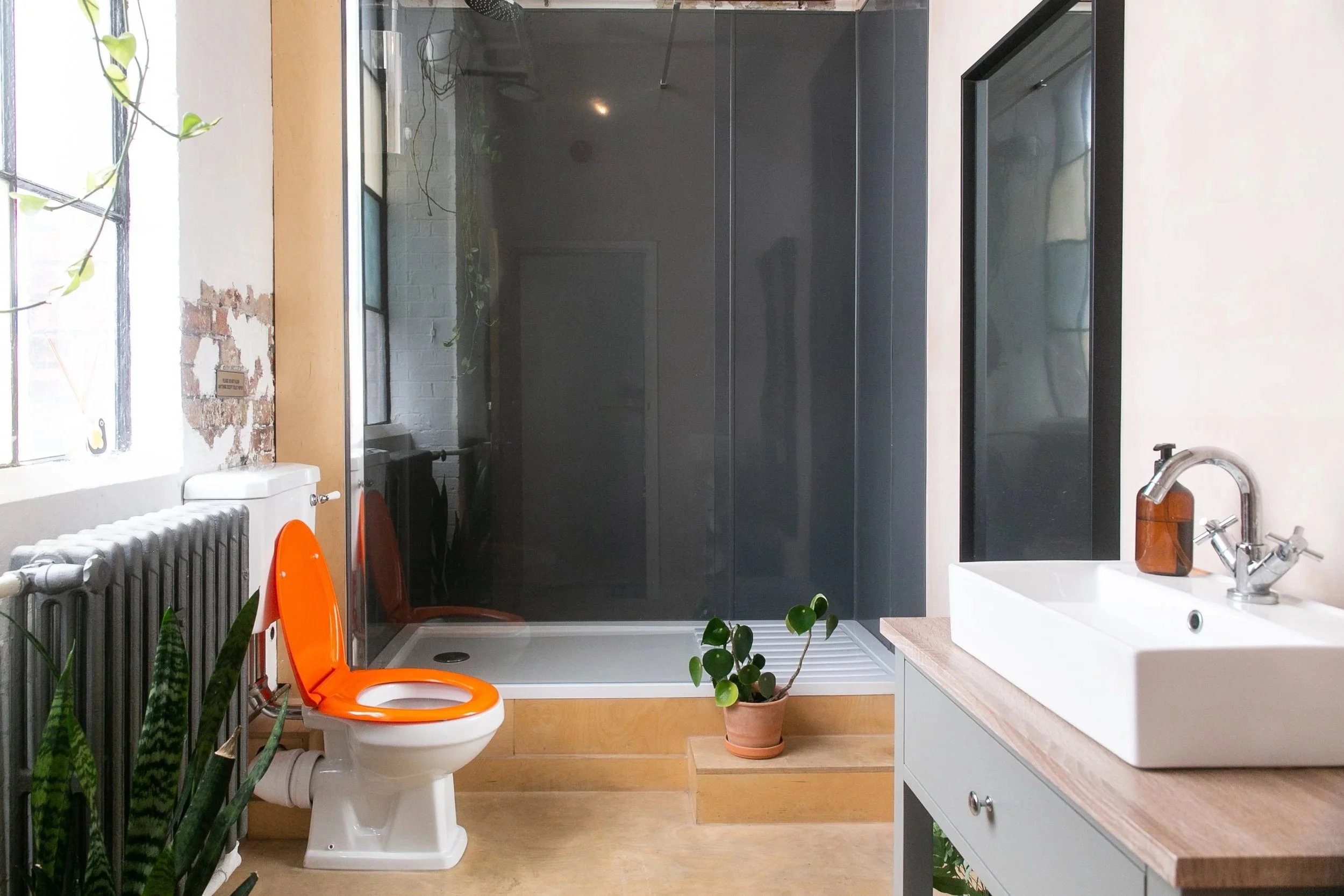 Modern bathroom with a white sink on a wooden vanity, orange toilet with open lid, large black shower enclosure, houseplant in a terracotta pot, and natural light from a window, with some exposed brick wall details.