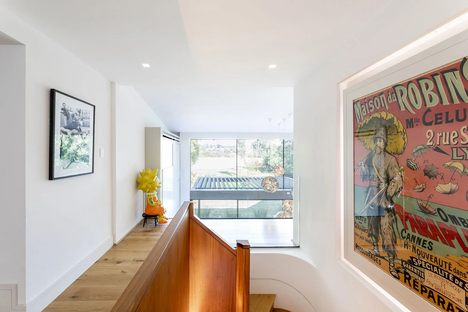 Modern indoor space with white walls, wooden flooring, large window, and decorative artwork, including a framed black-and-white photo, a colorful poster, and a yellow character sculpture at the end of the hallway.
