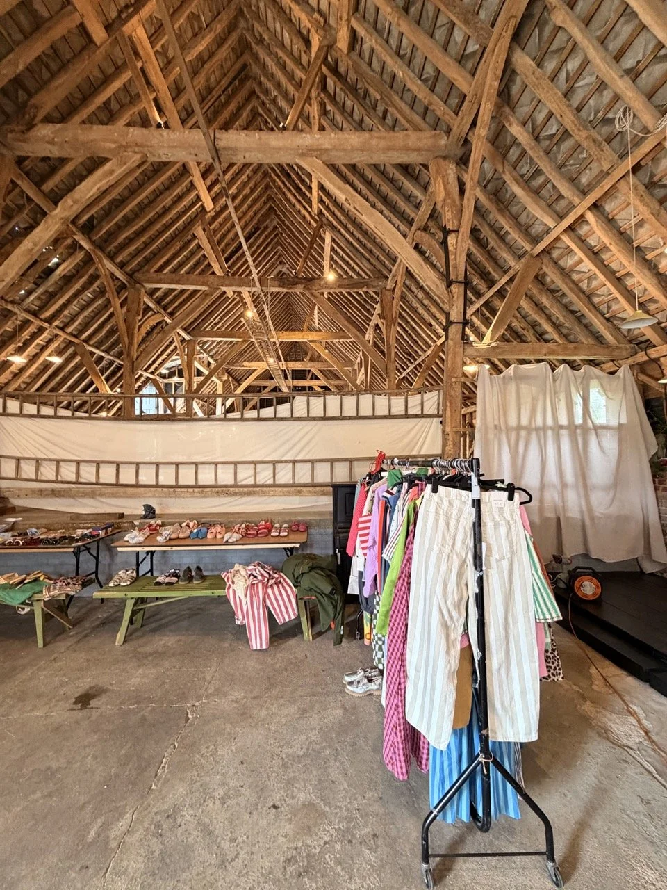 Indoor rummage sale setup in a barn with clothing rack, tables of shoes, sandals, and other items, and a wooden structure with exposed beams.