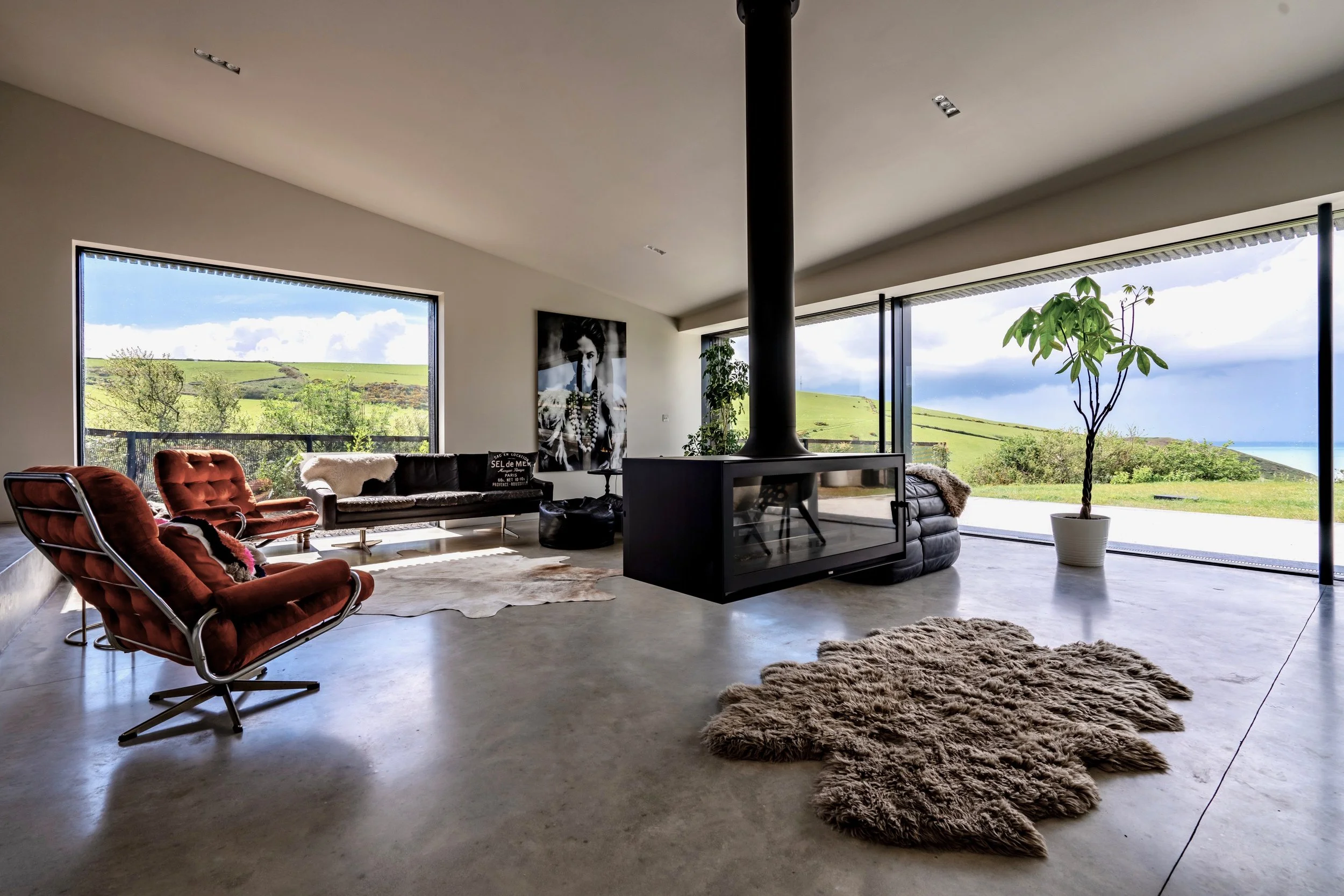 Modern living room with large windows showing green hills and blue sky, featuring red velvet chairs, black sofa, white textured rug, and a potted plant.