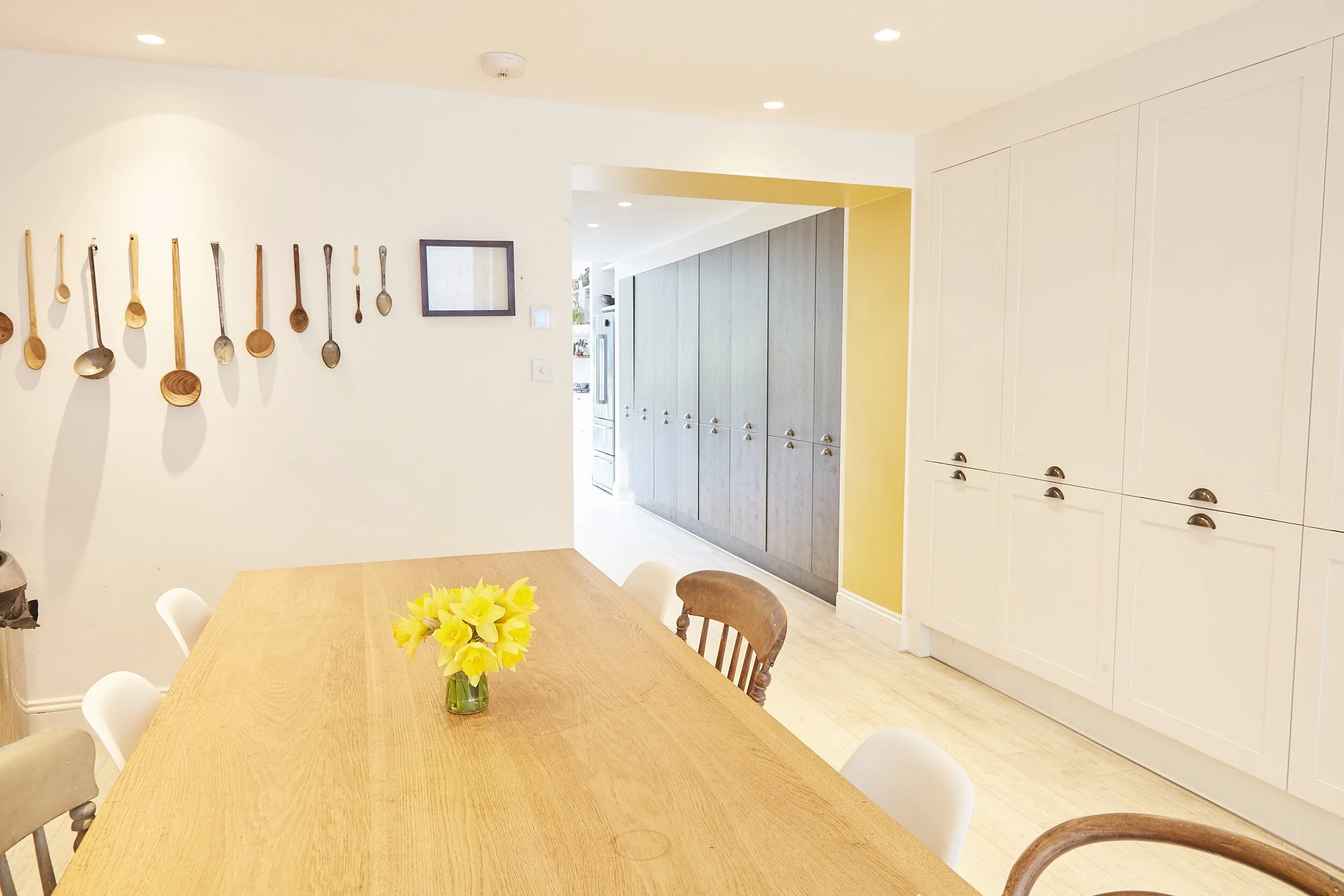 A dining room with a wooden table, a flower arrangement, and beige chairs. The wall features hanging wooden spoons and a small mirror. Beyond the doorway are gray cabinets and yellow walls.