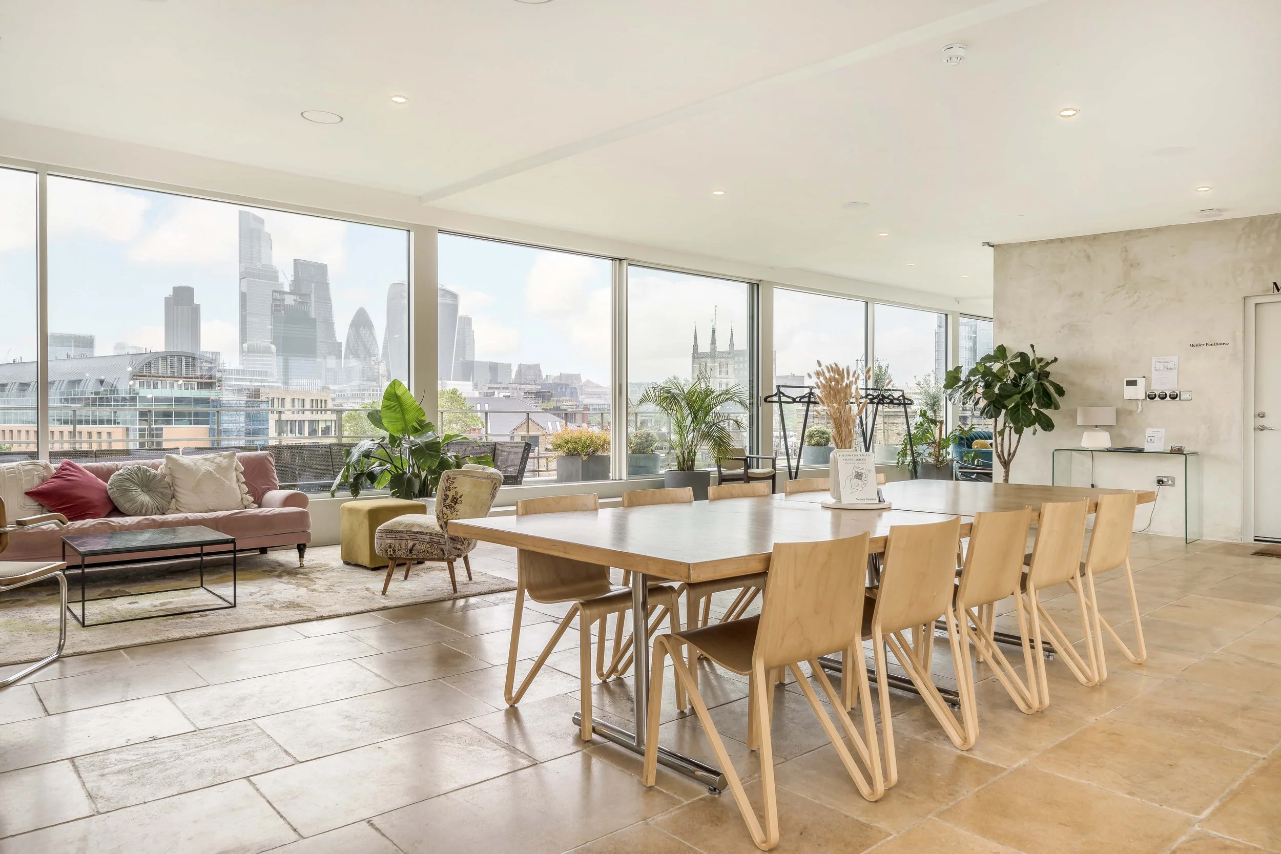 Modern lounge with large windows overlooking city skyline, pink sofa with pillows, wooden dining table with chairs, indoor plants, decorative items, and natural light.