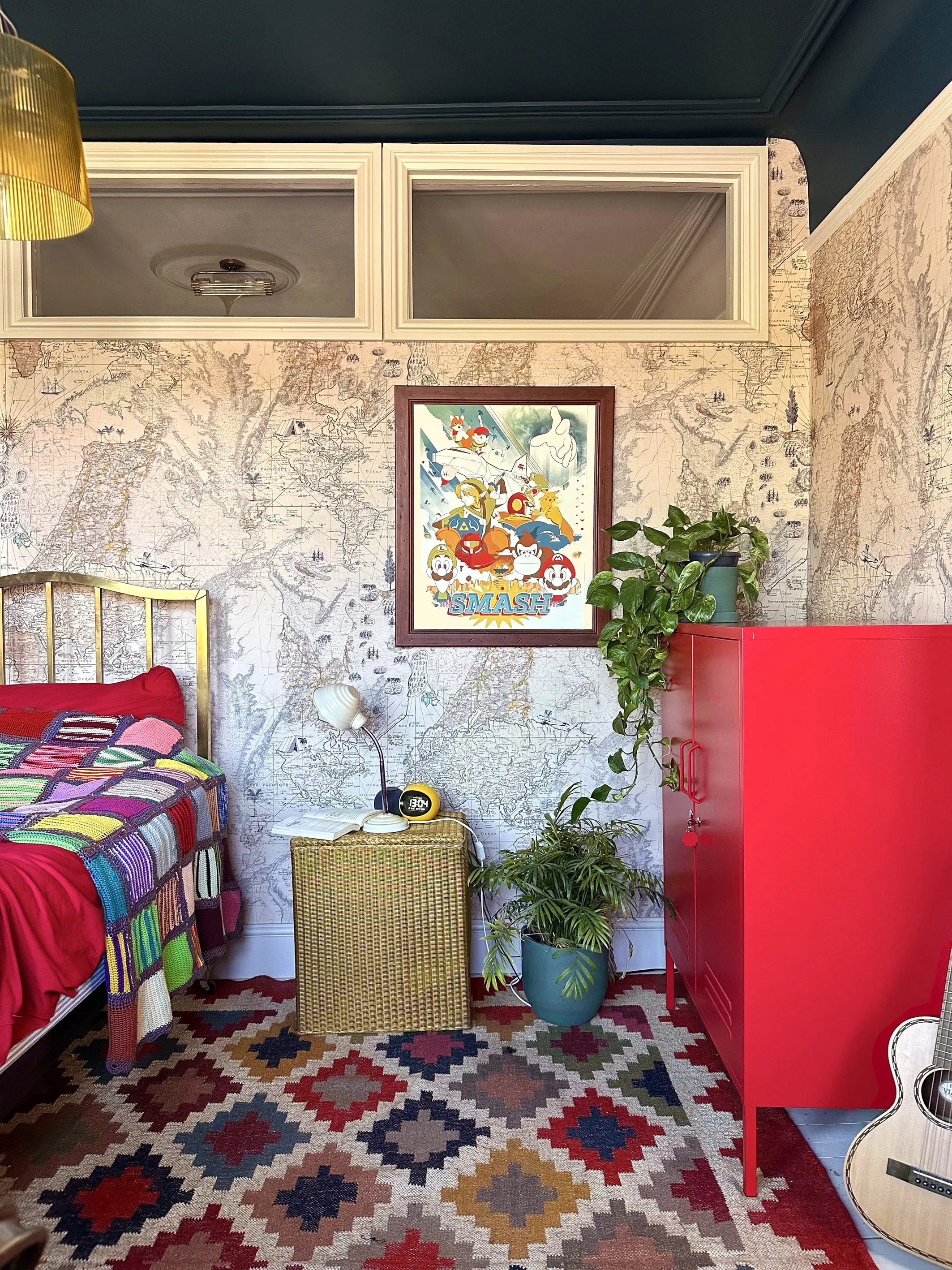 A bedroom with a colorful knitted blanket on the bed, a small bedside table with a lamp and an alarm clock, potted plants on a red cabinet, a framed art print on the wall, and a guitar on the floor. The wallpaper has a vintage map design, and the flo