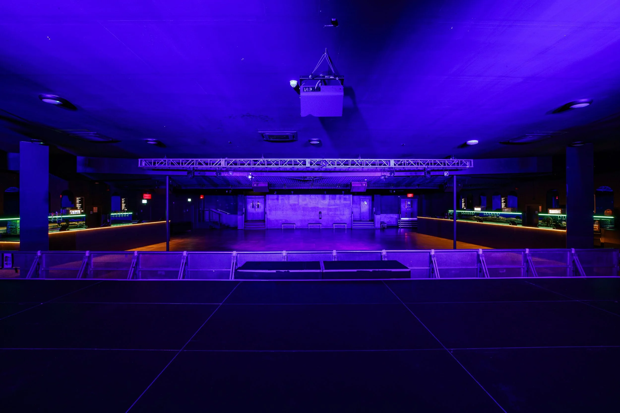 Empty nightclub with a dance floor illuminated by purple and green neon lights, with a stage and sound equipment at the back, and a bar area on the sides.