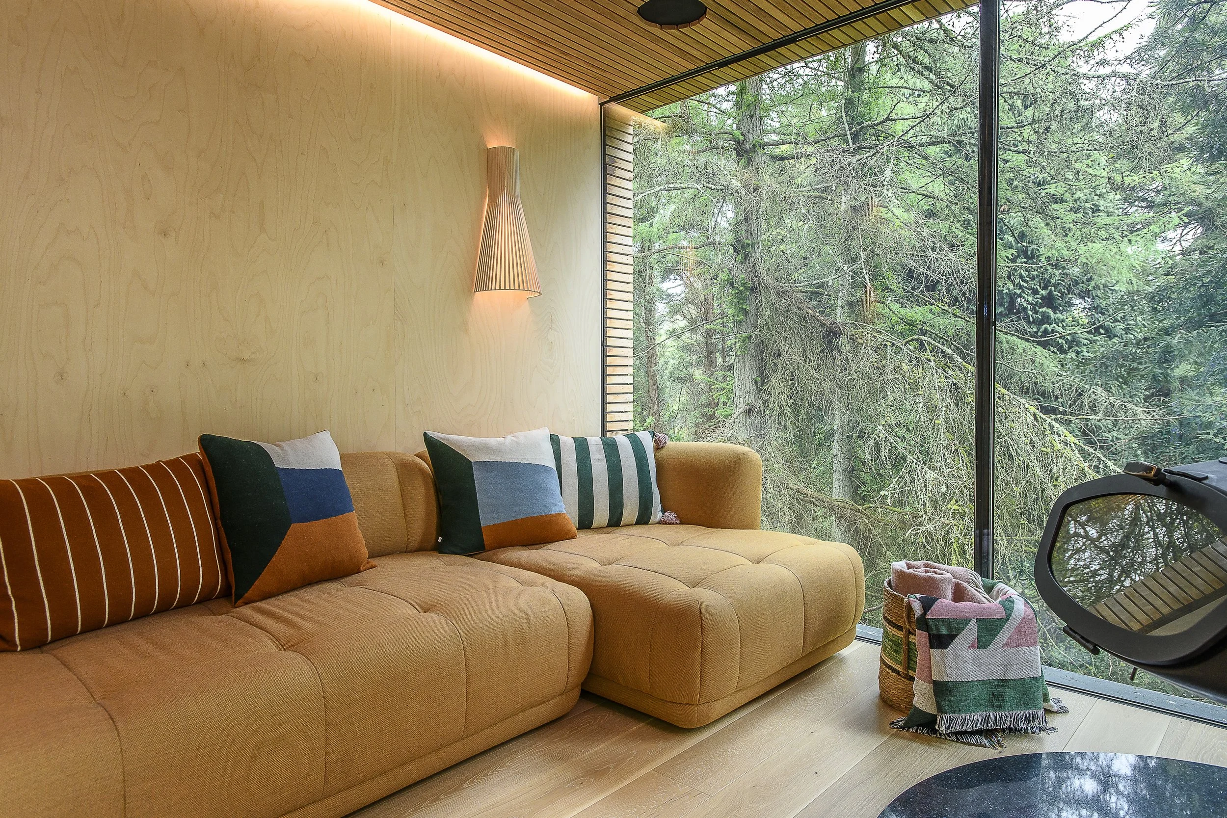 Living room with a tan sectional sofa, colorful striped pillows, wooden wall paneling, modern wall sconce, large glass window showing a forest view, woven baskets with textiles, and exercise equipment.