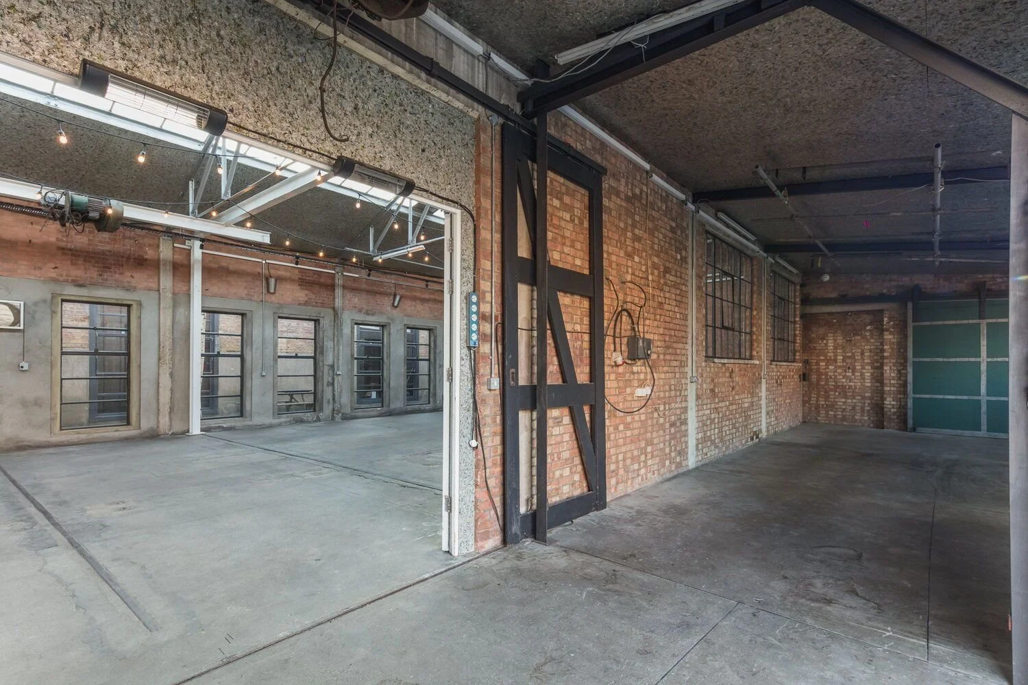 Interior view of an industrial-style brick building with large open spaces, exposed beams, and windows. There is a sliding metal door on the right, and the space appears to be a converted warehouse or studio with concrete floors and string lights han