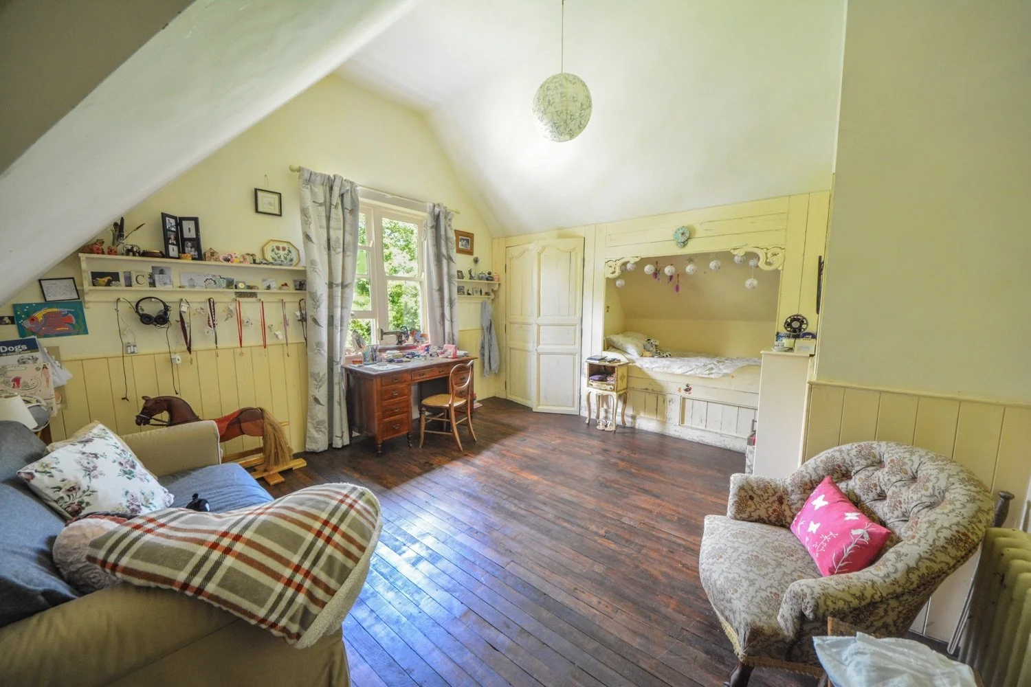 A cozy attic bedroom with yellow wooden-paneled walls, a window with patterned curtains, a desk, a sofa with floral pillows, an armchair with a pink pillow, and a bed built into the wall with hanging paper lanterns.
