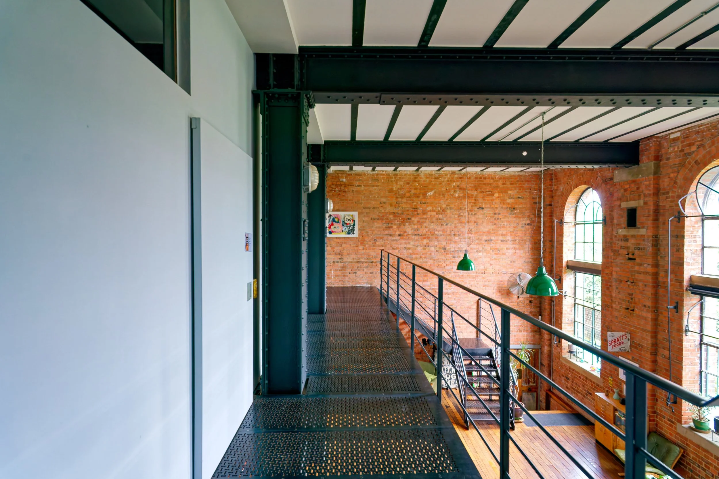 Interior view of a loft with exposed brick walls, large arched windows, green pendant lights, and a metal balcony with stairs.