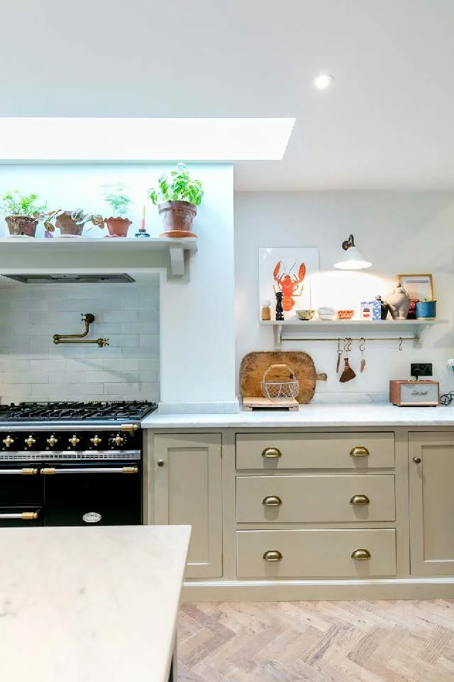 Kitchen with gray cabinets, a white countertop, and decorative shelves. Potted plants on a shelf above the stove, a framed artwork of a red animal face, a small lamp, and various kitchen items on the counters.