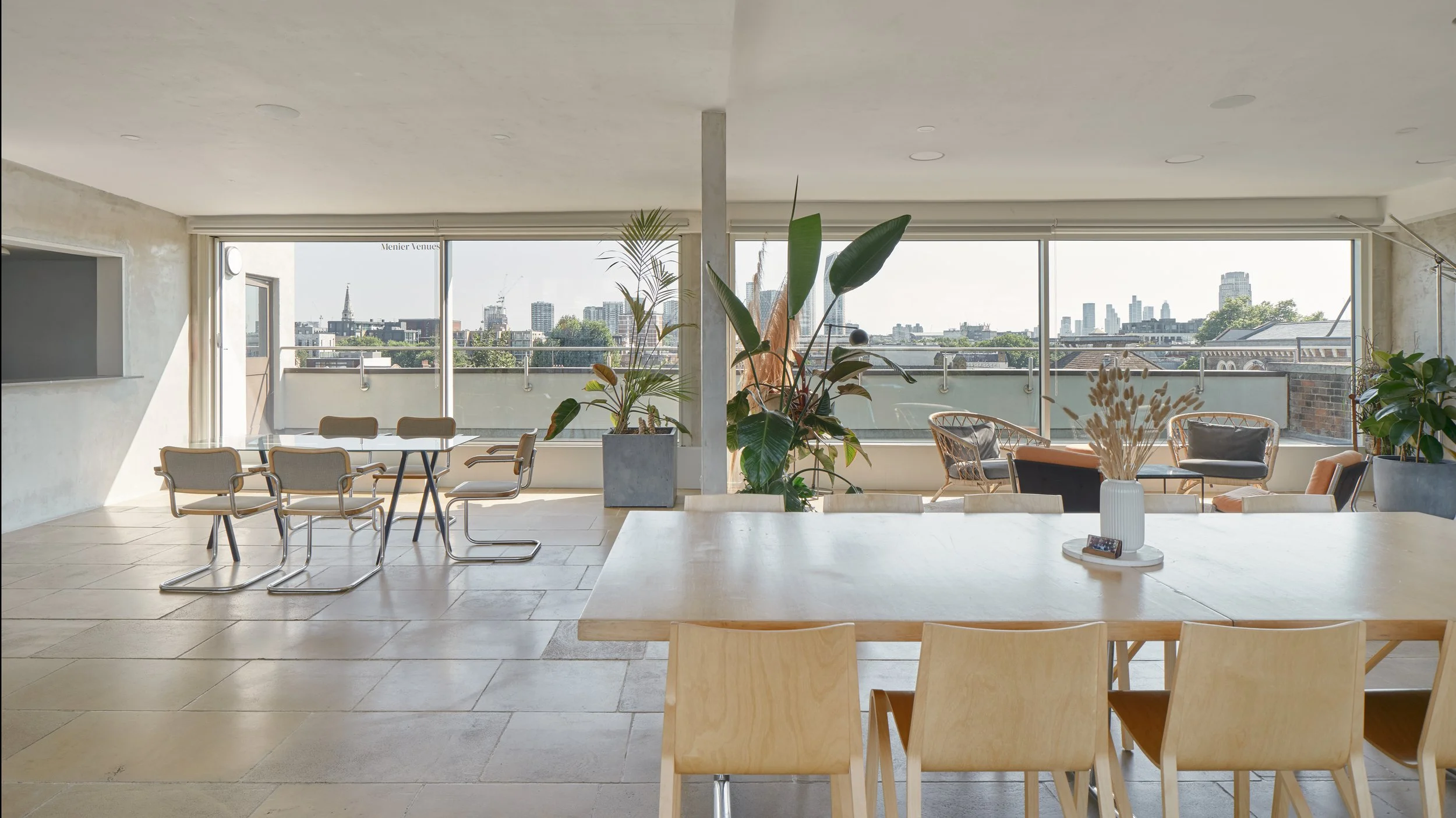 Bright, spacious office lounge or meeting area with large windows overlooking a cityscape, filled with natural light, wooden tables and chairs, potted plants, and modern decor.