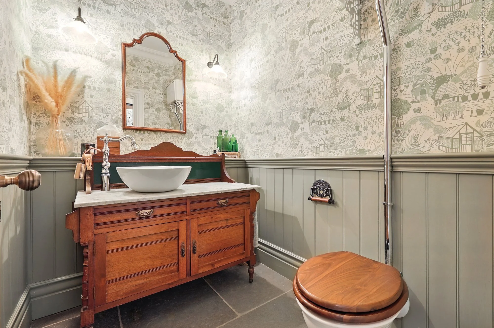 A vintage bathroom with wallpaper featuring village scenes, a wooden vanity with a vessel sink, a mirror, and a modern toilet with wooden seat lid.