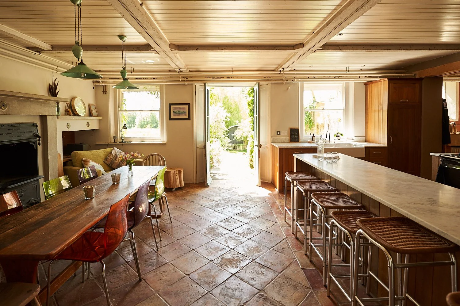 Bright kitchen with a large wooden dining table, colorful chairs, a kitchen island with bar stools, and open windows and doors letting in natural light.