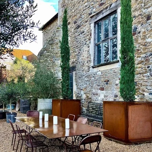 Outdoor patio with a wooden table, chairs, and candles in front of a stone building with large windows and tall green plants.