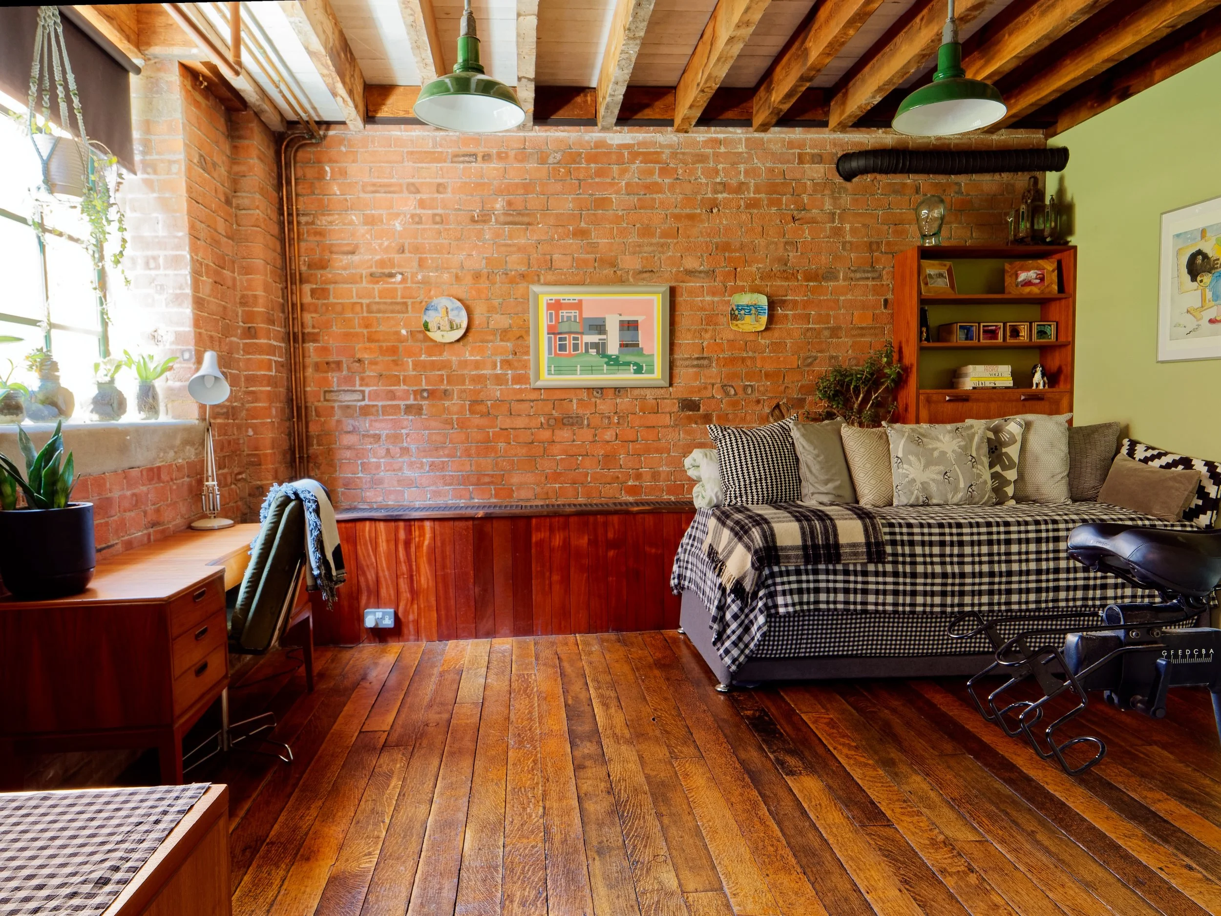 A cozy living room with a brick wall, wooden ceiling beams, and hardwood floors. Features a large sofa with various patterned cushions, a bookshelf with decorations and books, a small desk with plants and a lamp, and a window with sunlight streaming 