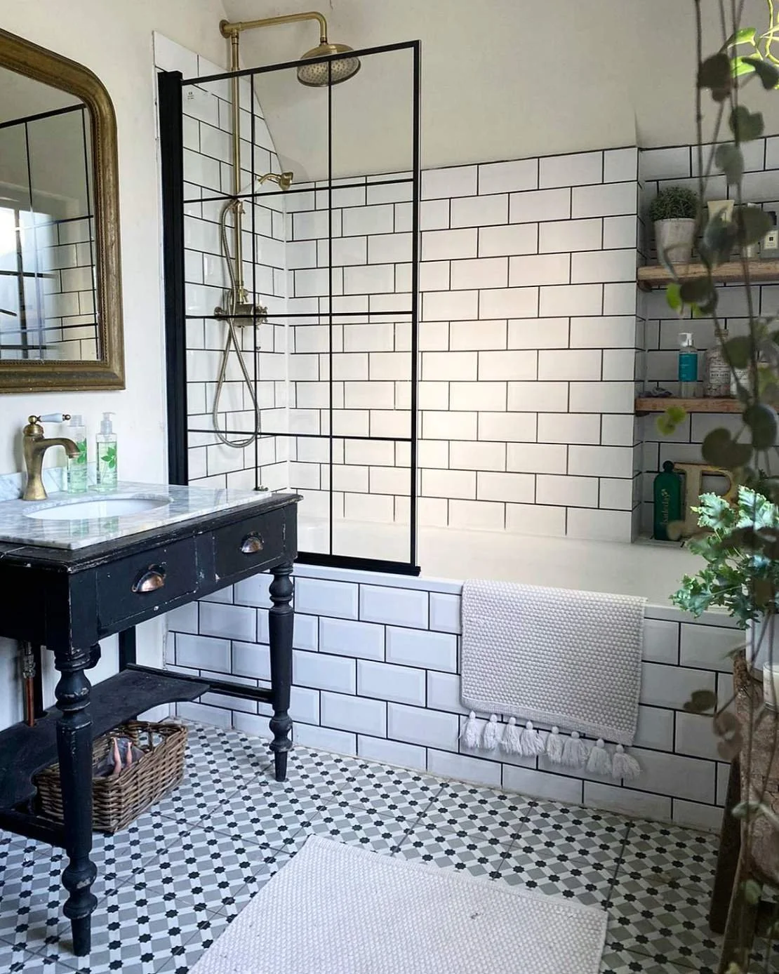 Bathroom with black-framed glass shower enclosure, wall tiles, vintage black vanity with marble top, mirror, white textured towel, wooden shelf with plants and toiletries, patterned tile floor, and a white rug.