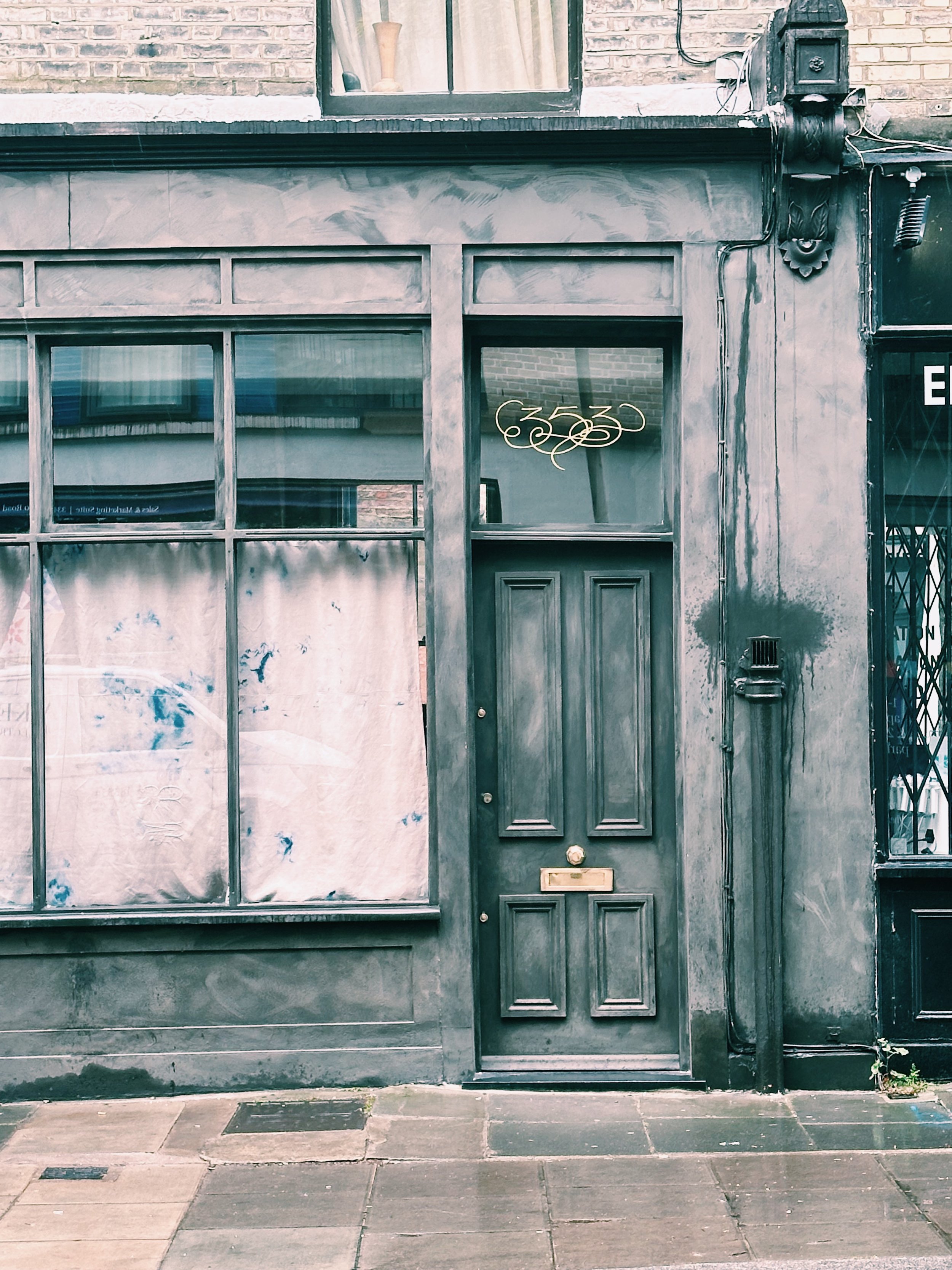 Gray building facade with large window pane and wooden door, neon sign in the window, and some electrical wiring and fixtures on the exterior wall.
