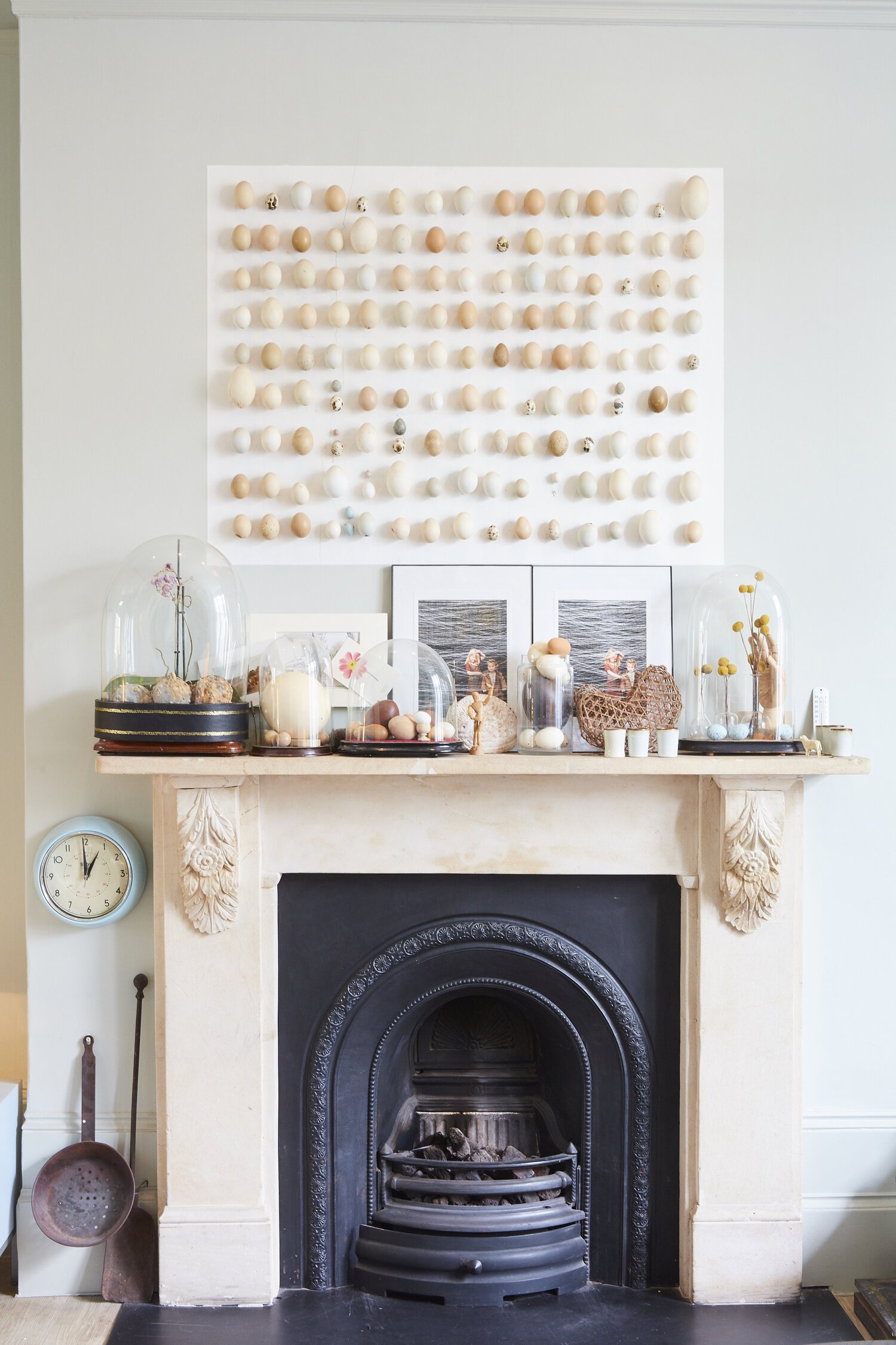 Decorative shelf above a fireplace with various glass domes containing rocks, eggs, and flowers, and framed black-and-white photographs, against a white wall with a circle clock nearby.
