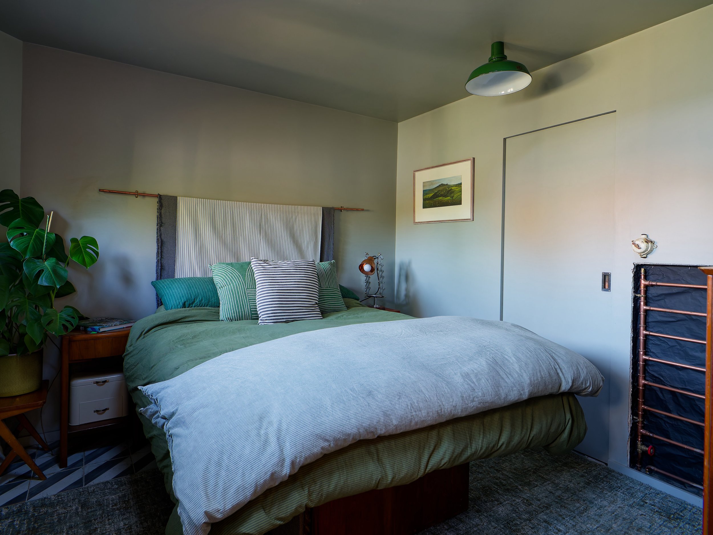 A cozy bedroom with a bed dressed in green and beige bedding, multiple pillows, a side table with a plant, artwork on the wall, and a ceiling light fixture.