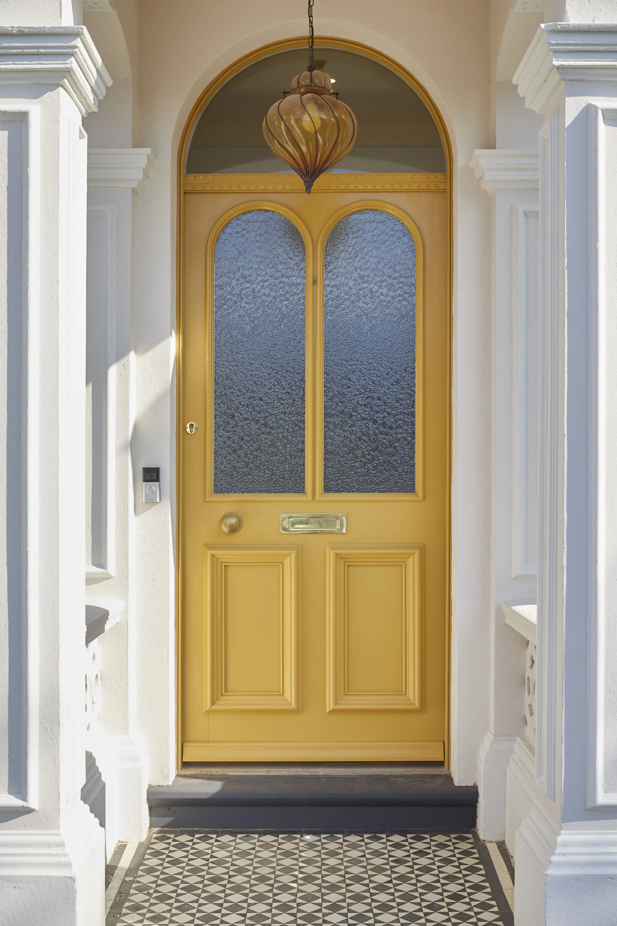 Yellow front door with frosted glass panels, surrounded by white columns and walls, with a decorative hanging lantern light above and a patterned tile entryway.