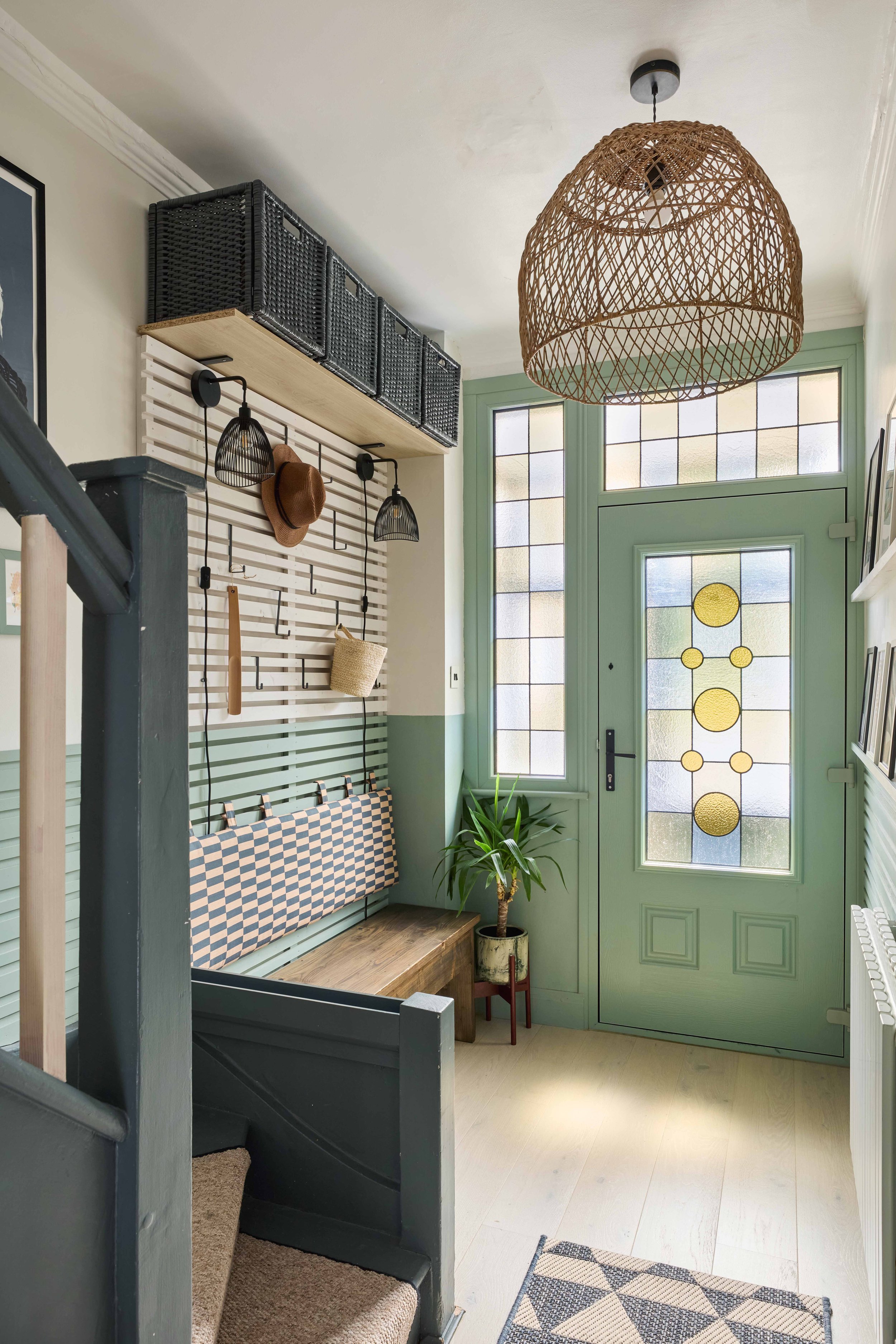 Entryway with green door and stained glass window, wooden bench, potted plant, woven pendant light, wall hooks with hats and bags, above storage baskets, and a striped door panel.