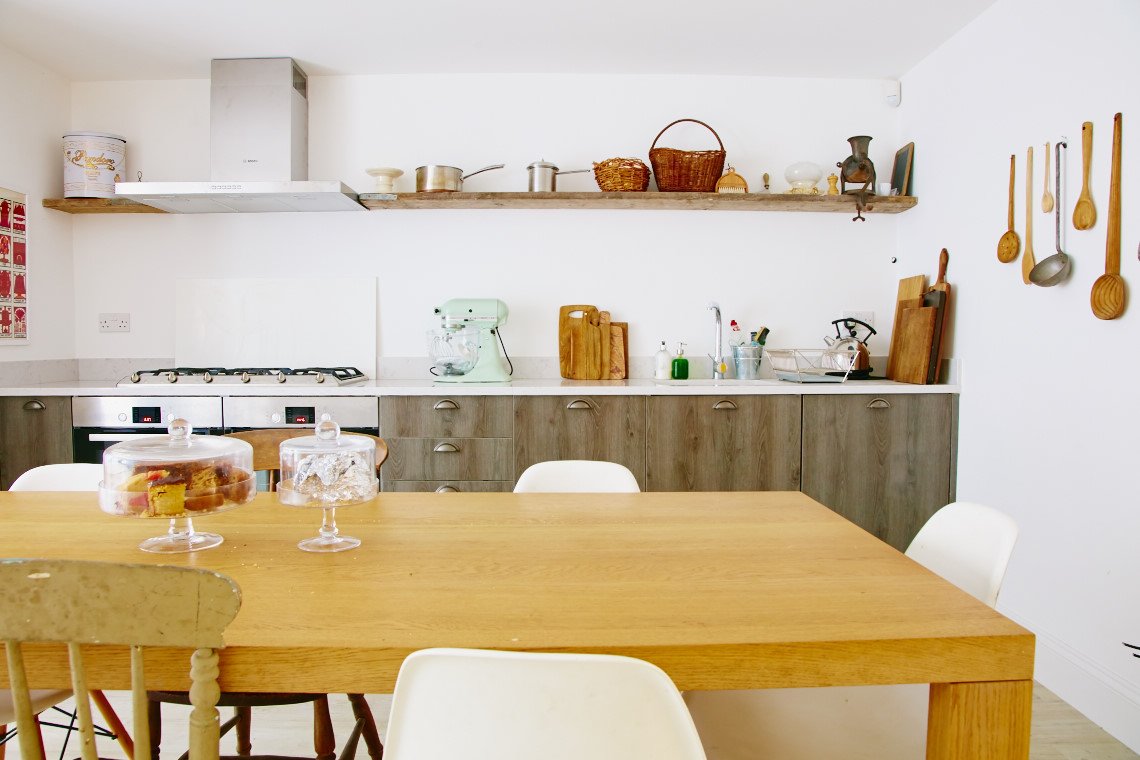 Kitchen with a wooden dining table and white chairs, open shelves with baskets and kitchen utensils, and a countertop with appliances and cutting boards.