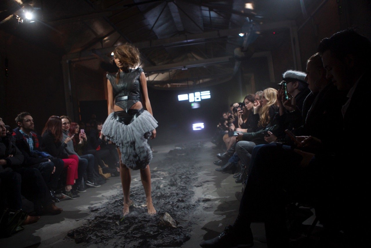 Model walking barefoot on a runway covered in ash during a fashion show, audience members taking photos and videos.
