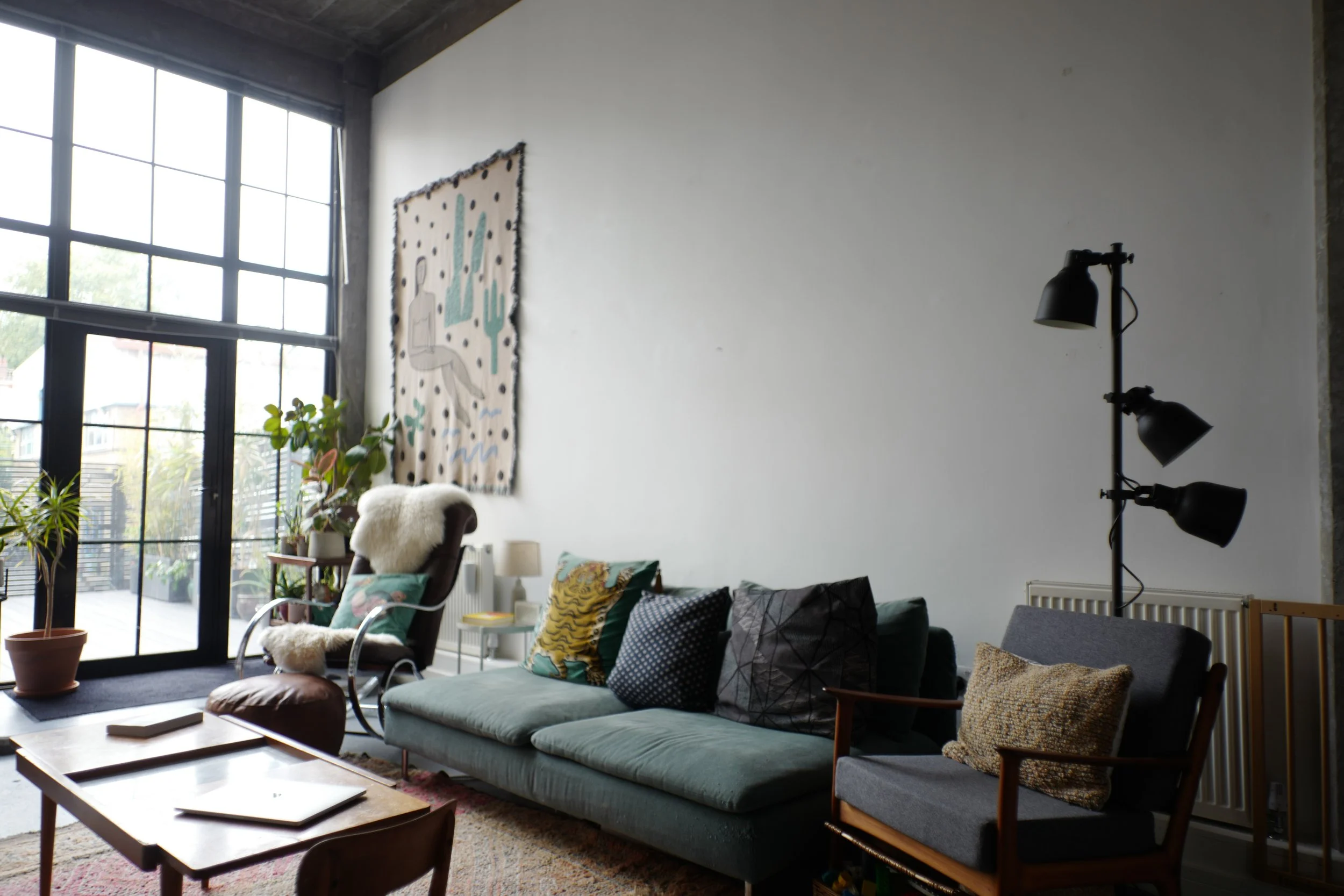 Living room with large windows, a green sofa with multiple cushions, a black armchair with a pillow, a tall potted plant, a floor lamp, a wall hanging art, and a coffee table.