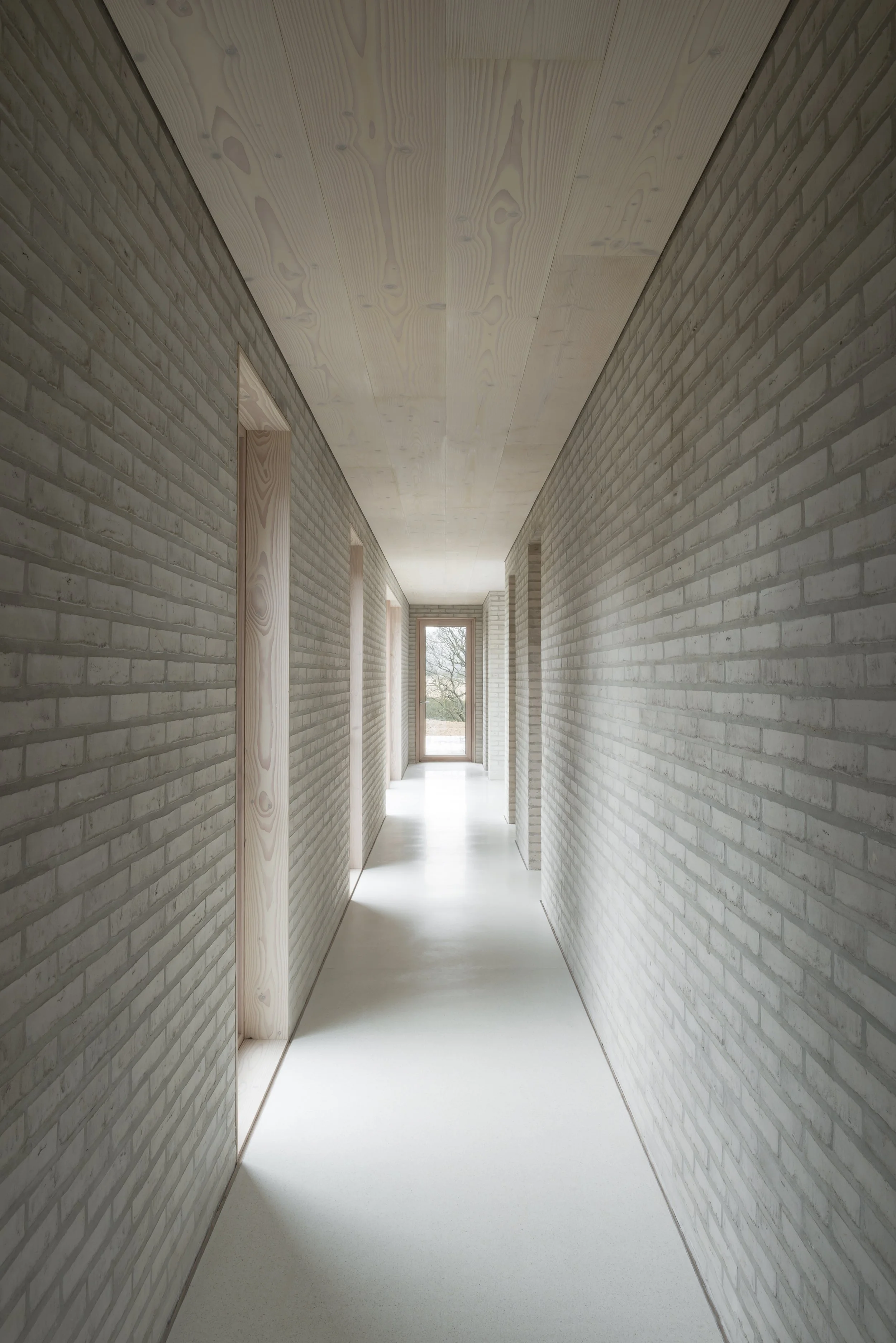 Minimalist indoor corridor with brick walls, light wood ceiling, and white floor leading to a glass door with a view of trees outside.