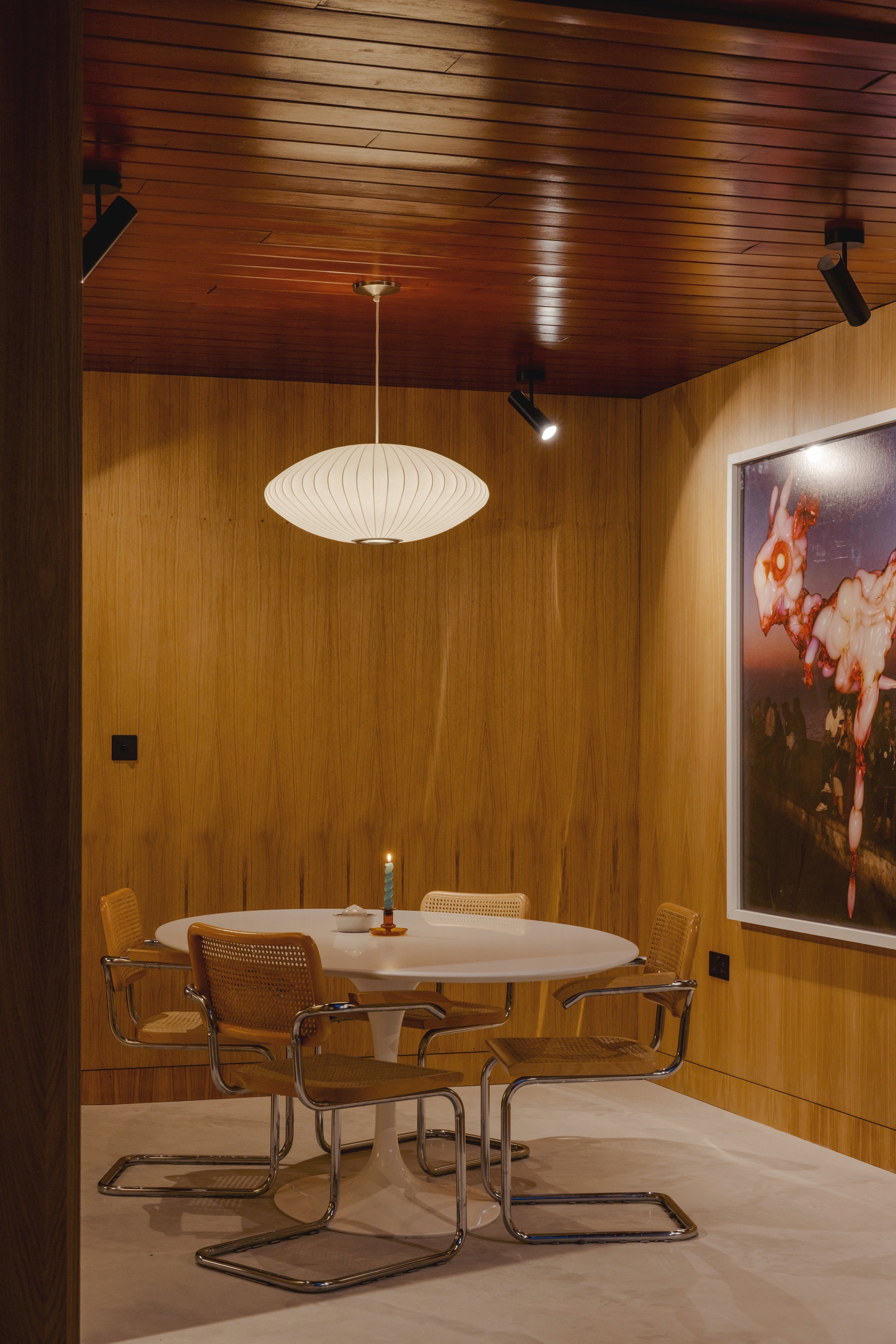 A cozy dining area with a round white table, four wooden and metal chairs, a blue birthday candle on a small cake, a white paper lantern hanging from the wooden ceiling, a modern artwork on the wooden-paneled wall, and warm lighting.